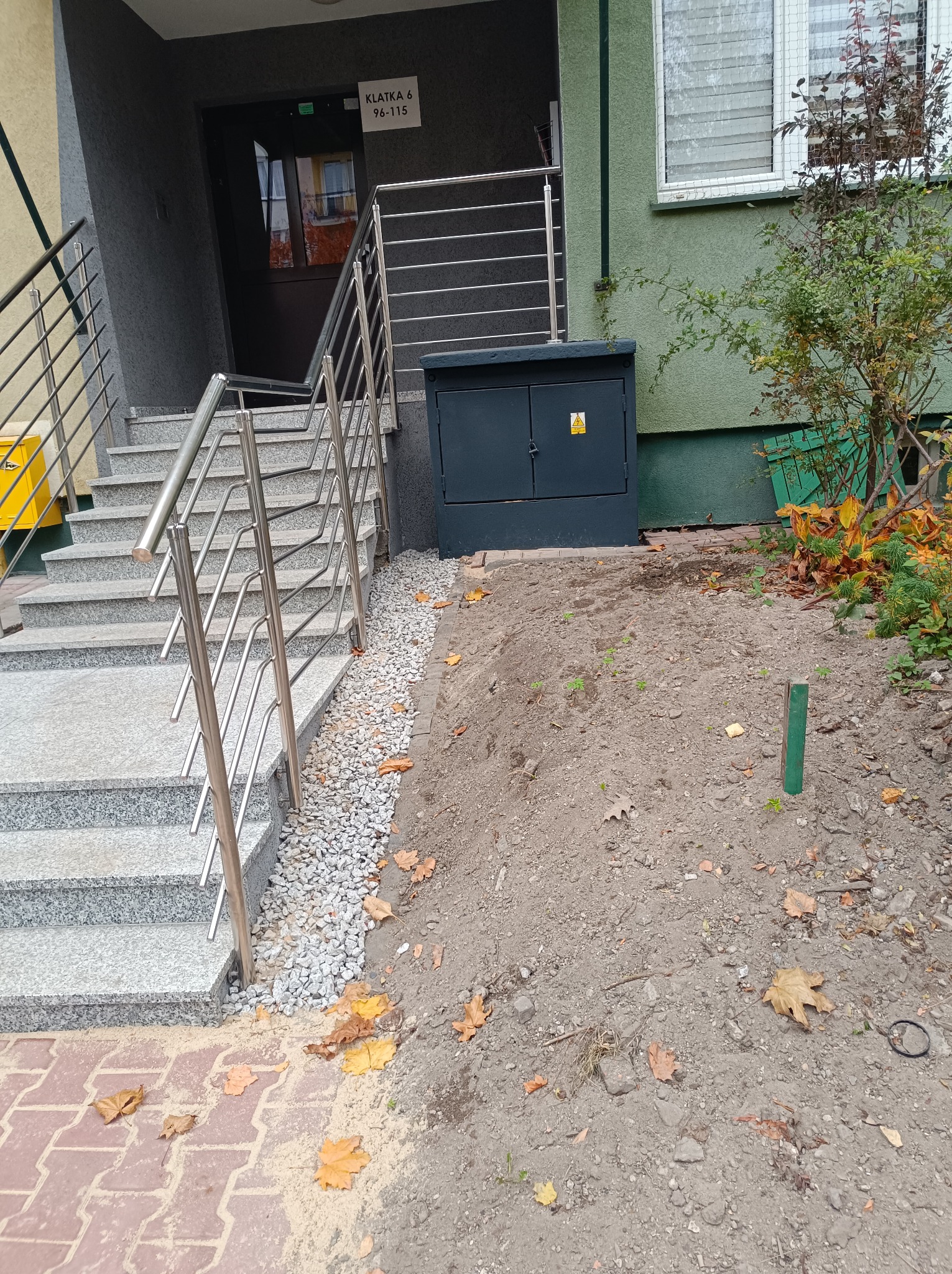 Wejście do klatki schodowej z granitowymi schodami i metalową barierką, obok skrzynka elektryczna, jesienne liście na kostce i ziemi.