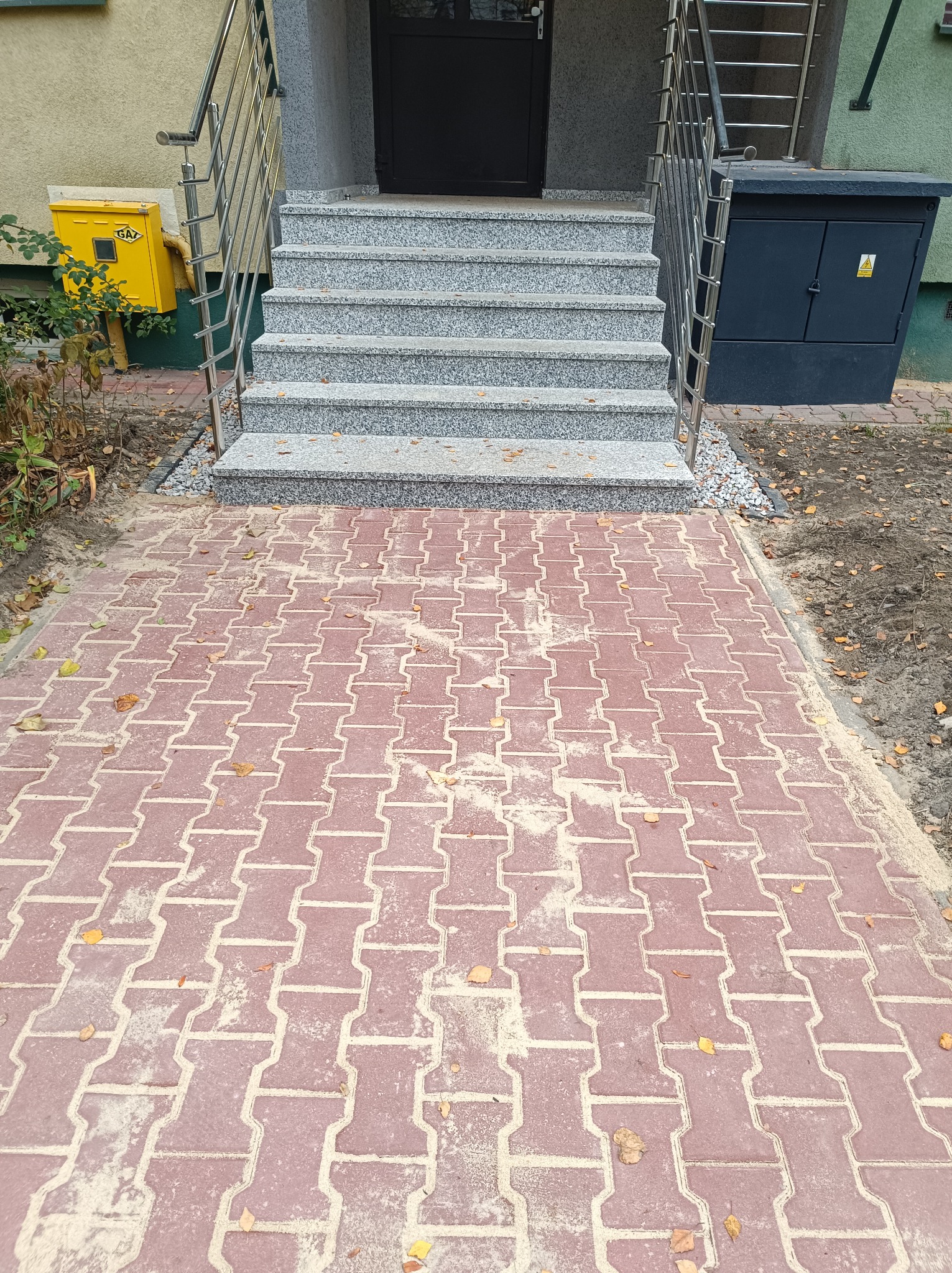 Czerwona kostka brukowa ułożona przed schodami wejściowymi do budynku, z żółtą skrzynką gazową i szarą szafką elektryczną po bokach.