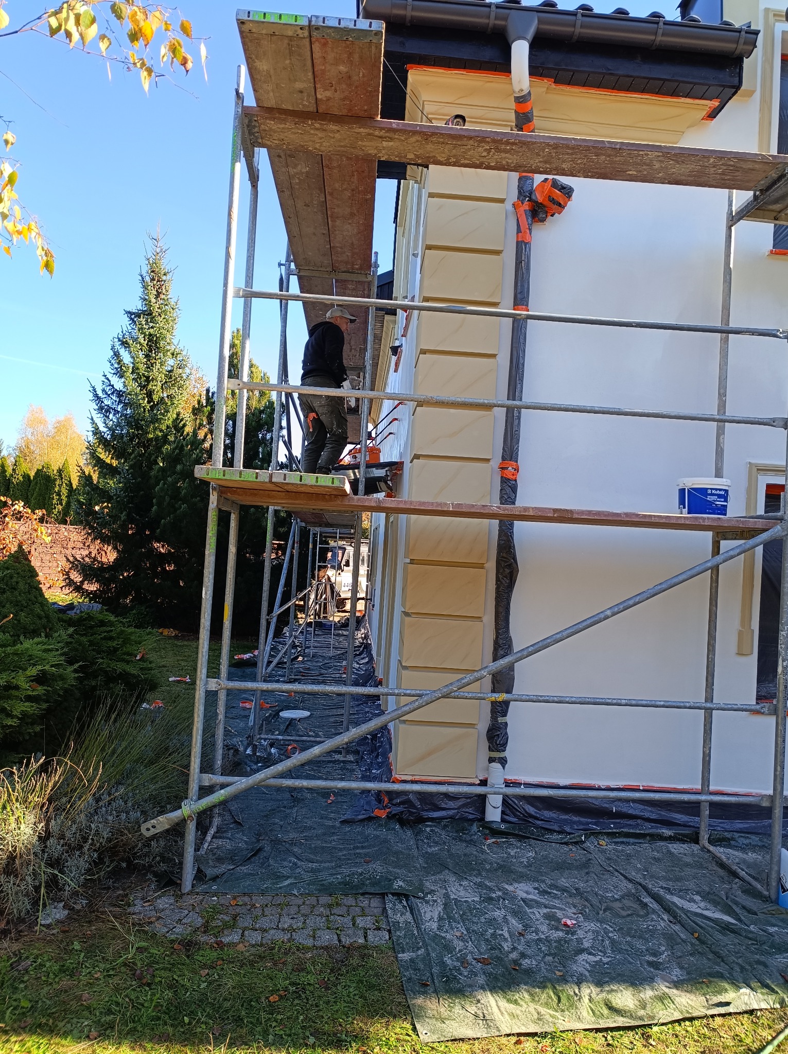 Pracownik na rusztowaniu maluje elewację domu z dekoracyjnymi elementami, zabezpieczoną folią, widoczna puszka z farbą, w tle drzewa i błękitne niebo.