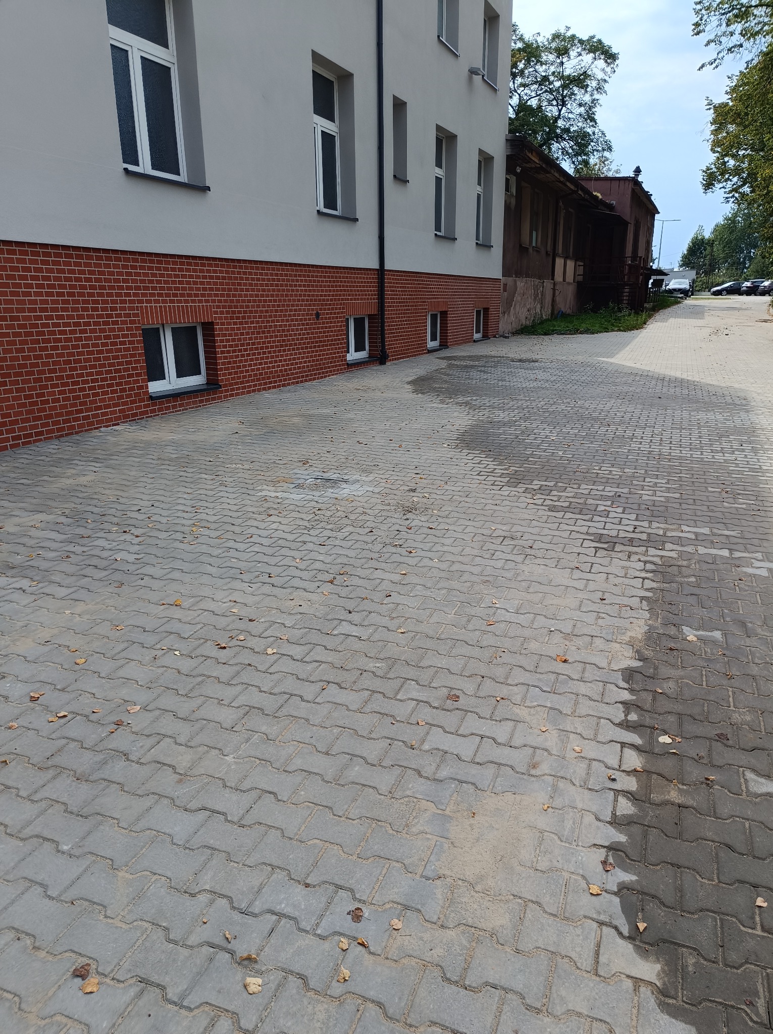 Szara kostka brukowa ułożona przed budynkiem z ceglaną podmurówką i szarą elewacją, widoczne liście i mokre plamy na kostce.
