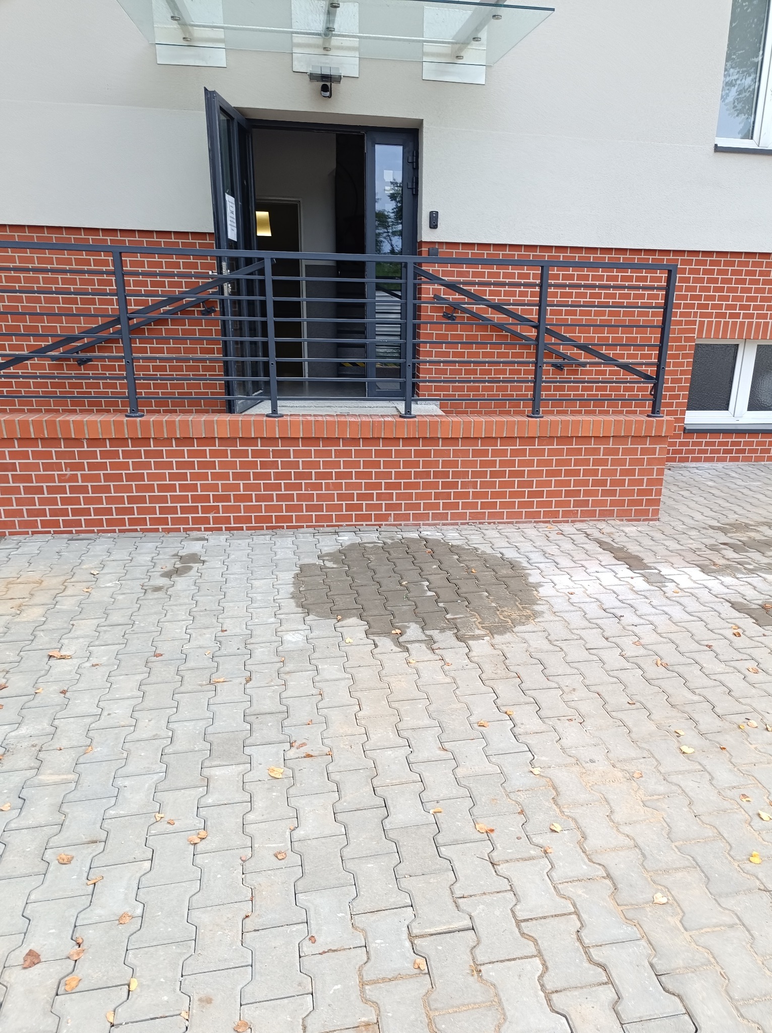Wejście do budynku z otwartymi drzwiami, ceglanym podwyższeniem, czarną balustradą i szarą kostką brukową na zewnątrz, z widocznymi plamami wody na kostce.