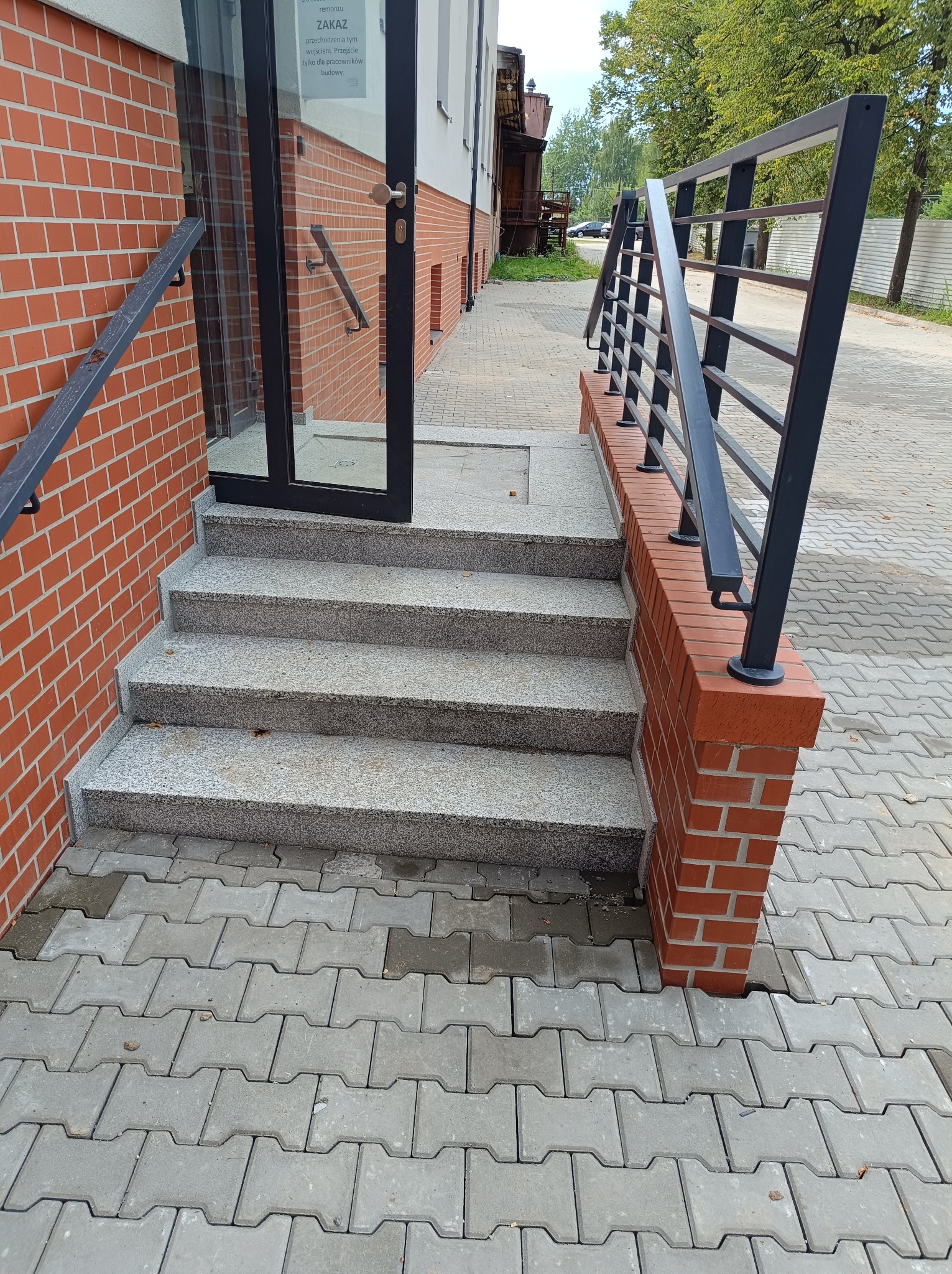 Granitowe schody wejściowe z czarną metalową balustradą prowadzące do budynku z ceglaną elewacją i szklanymi drzwiami, wykończone szarą kostką brukową.