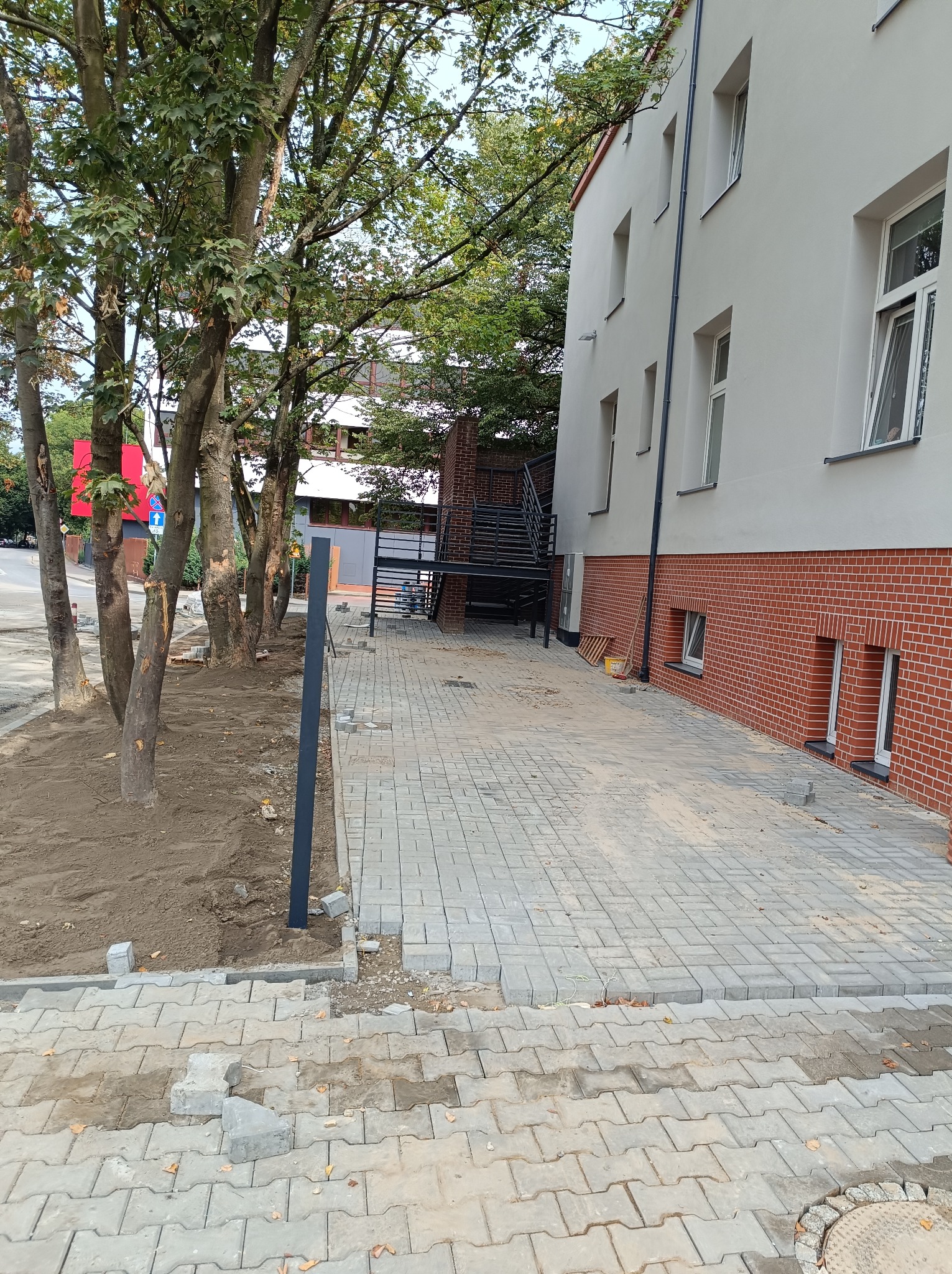 Układanie szarej kostki brukowej na chodniku przy budynku z ceglaną podmurówką, widoczne drzewa i przygotowany teren pod nasadzenia.