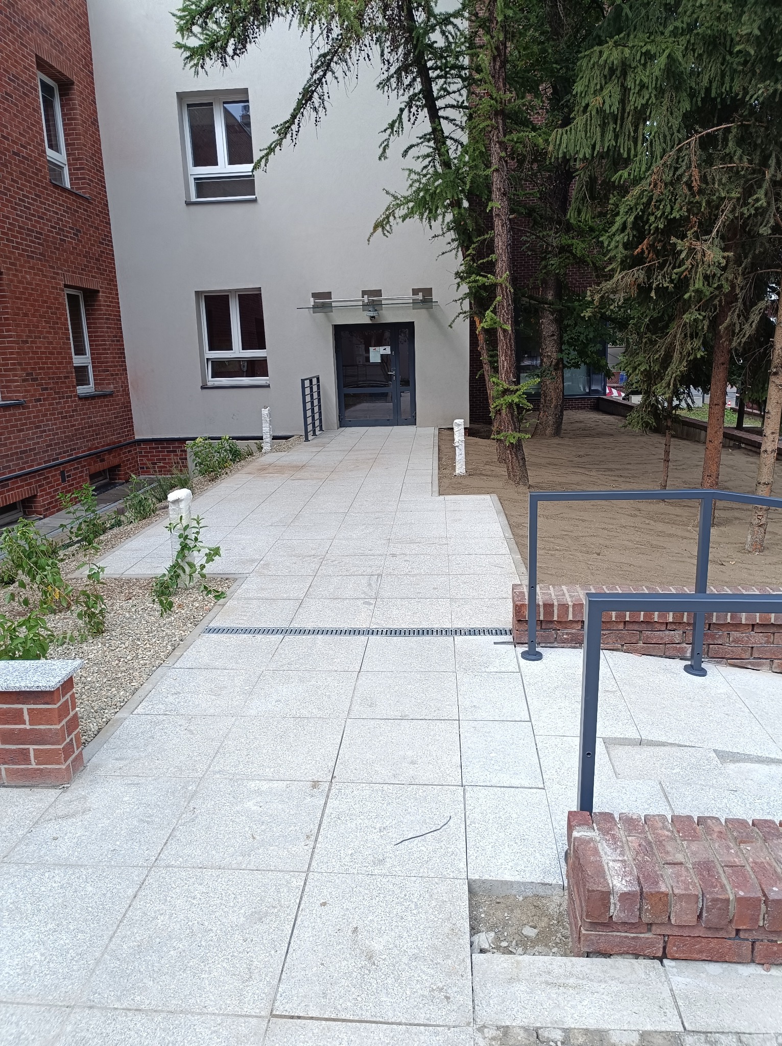 Jasnoszara kostka brukowa prowadząca do wejścia budynku, widoczne obrzeża z cegły, zieleń i elementy małej architektury.