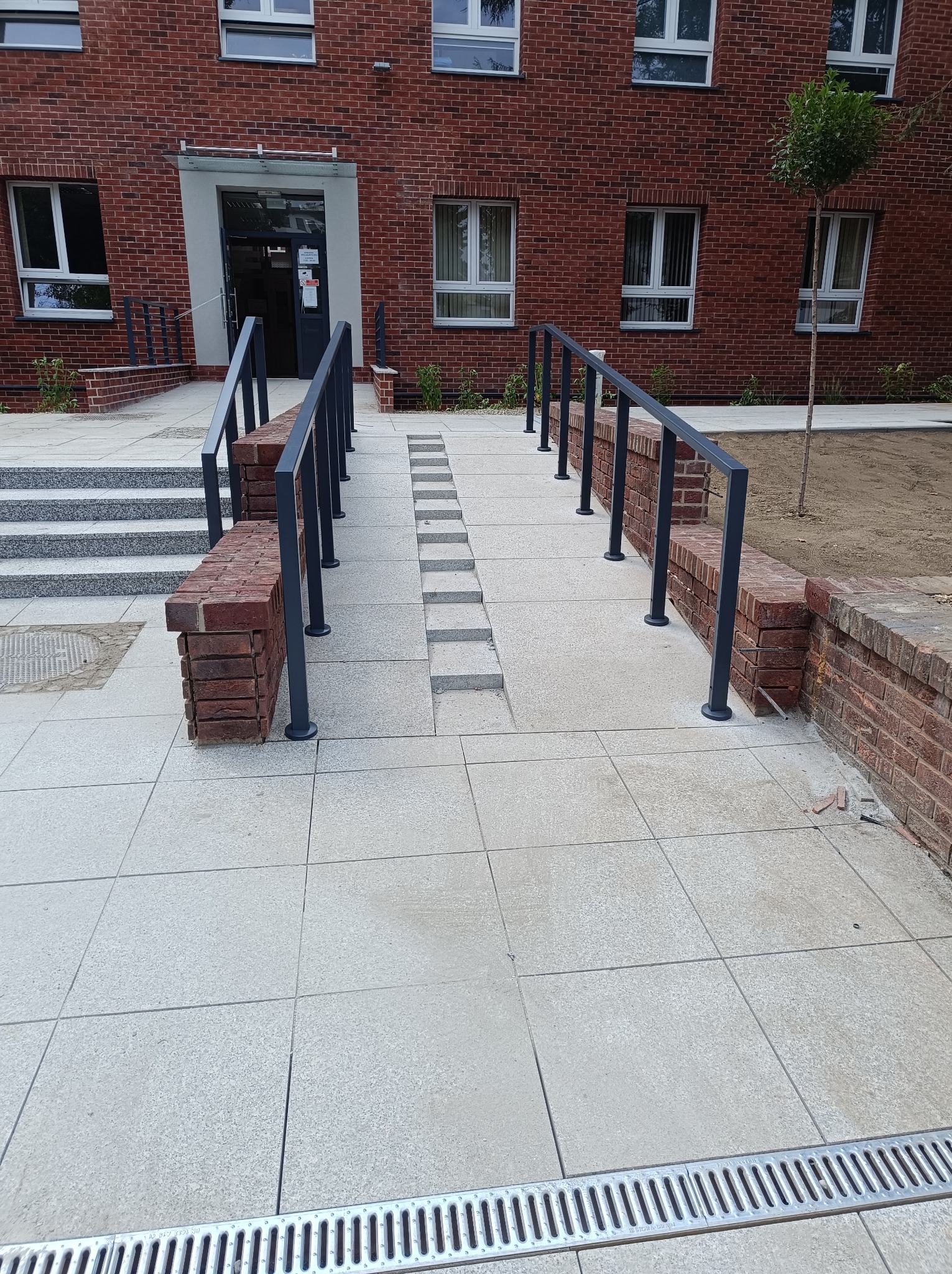 Nowe podejście do budynku z cegły z rampą dla wózków inwalidzkich i schodami, wykończone jasną kostką brukową oraz metalowymi barierkami z ceglanymi podstawami. Widoczne są także kratki odpływowe.