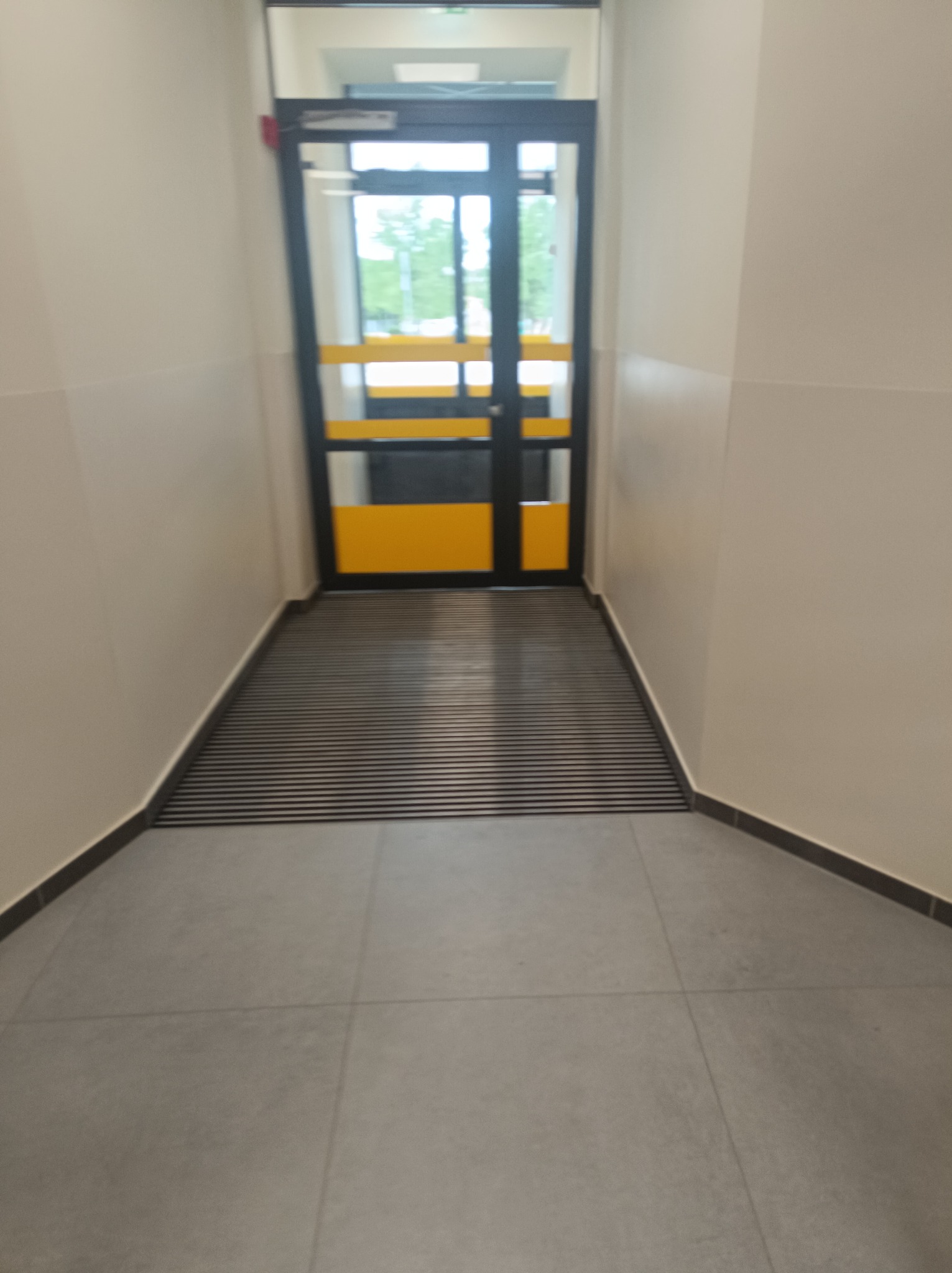 Wnętrze korytarza z szarą podłogą, żółto-czarnymi drzwiami i matą wejściową z poziomych listew.