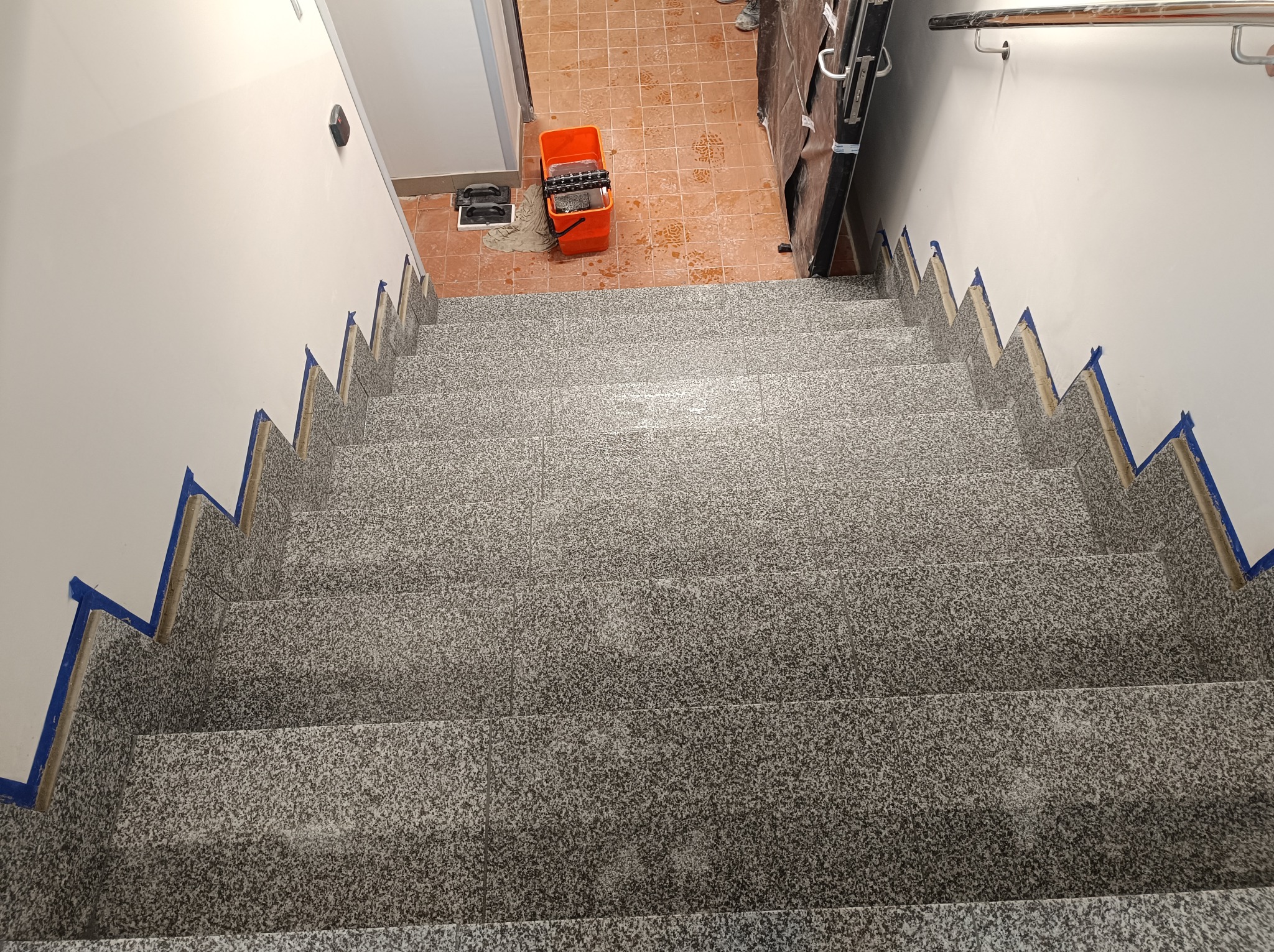 Widok z góry na schody pokryte szarymi płytkami imitującymi kamień, zabezpieczone niebieską taśmą malarską przy ścianach, z widocznym wiadrem z narzędziami malarskimi i śladami stóp na podłodze w tle.