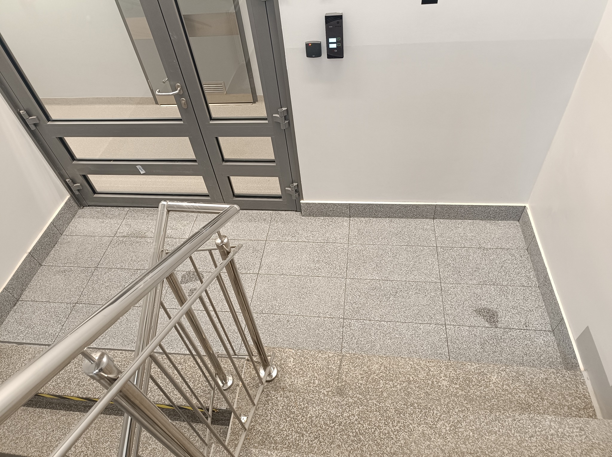 Wnętrze klatki schodowej z szarymi płytkami podłogowymi, metalową balustradą i szklanymi drzwiami wejściowymi z szarą ramą, widok z góry.