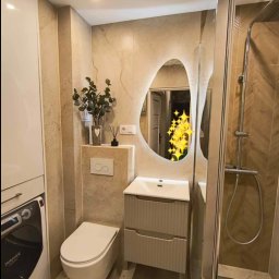 Tomek Sa - Nowoczesna łazienka z podświetlanym lustrem o nieregularnym kształcie, kabiną prysznicową typu walk-in z drewnopodobnymi płytkami ułożonymi w jodełkę, wiszącą toaletą i pralką w zabudowie.