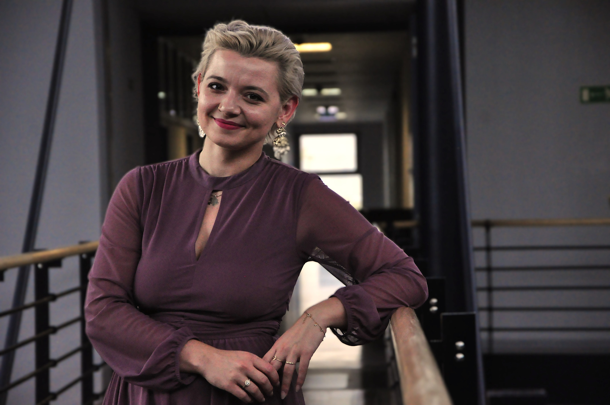 Uśmiechnięta blondynka w fioletowej sukience, oparta o balustradę w nowoczesnym biurze, z widocznymi kolczykami i pierścionkami.