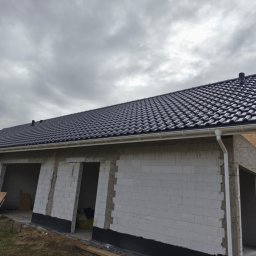 Daniel Wołoszyn - Nowy, czarny dach z rynnami na budynku w trakcie budowy, widok z dołu na tle pochmurnego nieba. Białe ściany z betonu komórkowego, widoczne otwory okienne i drzwiowe.