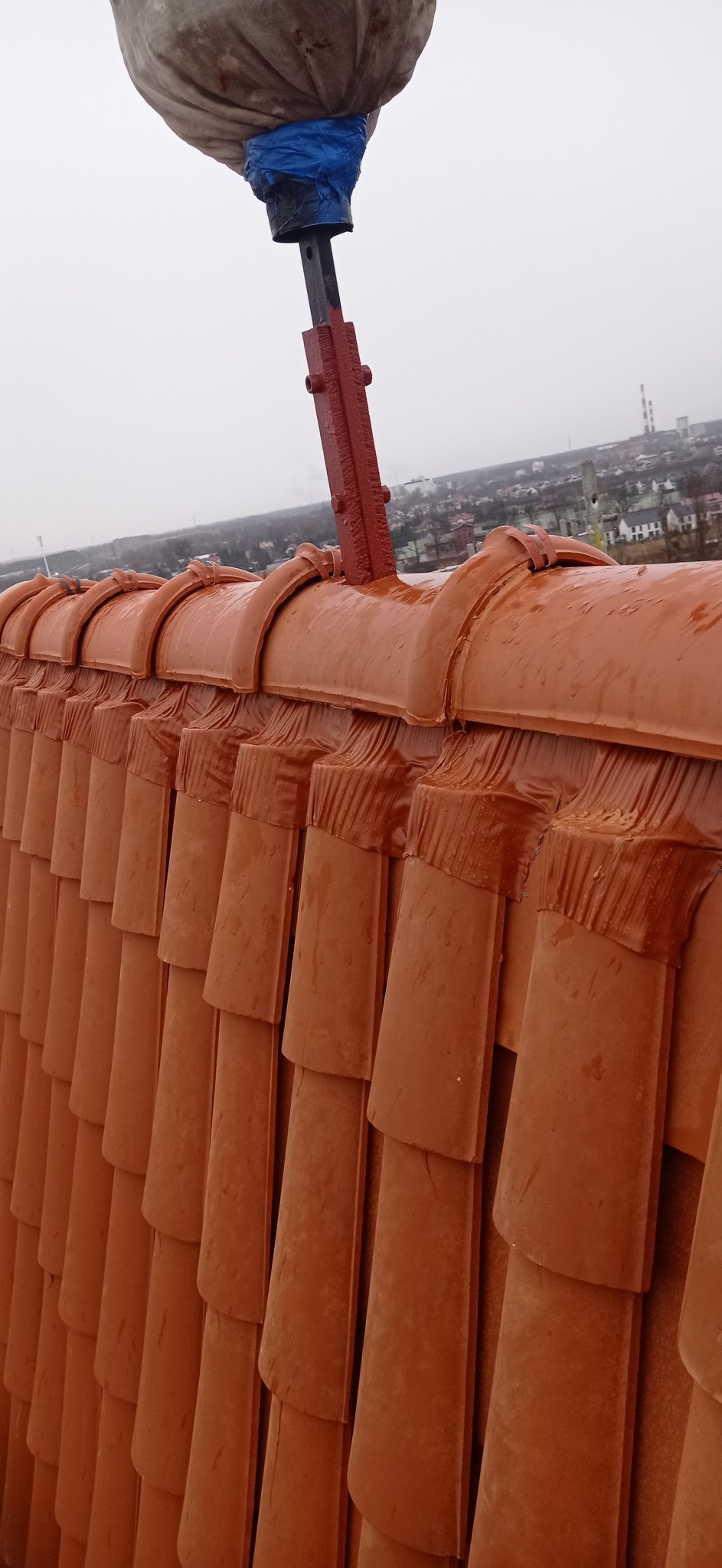 Czerwona dachówka ceramiczna, mokra od deszczu, z widocznym uchwytem narzędzia dekarskiego i workiem zabezpieczającym w tle, panorama miasta w mglistej perspektywie.