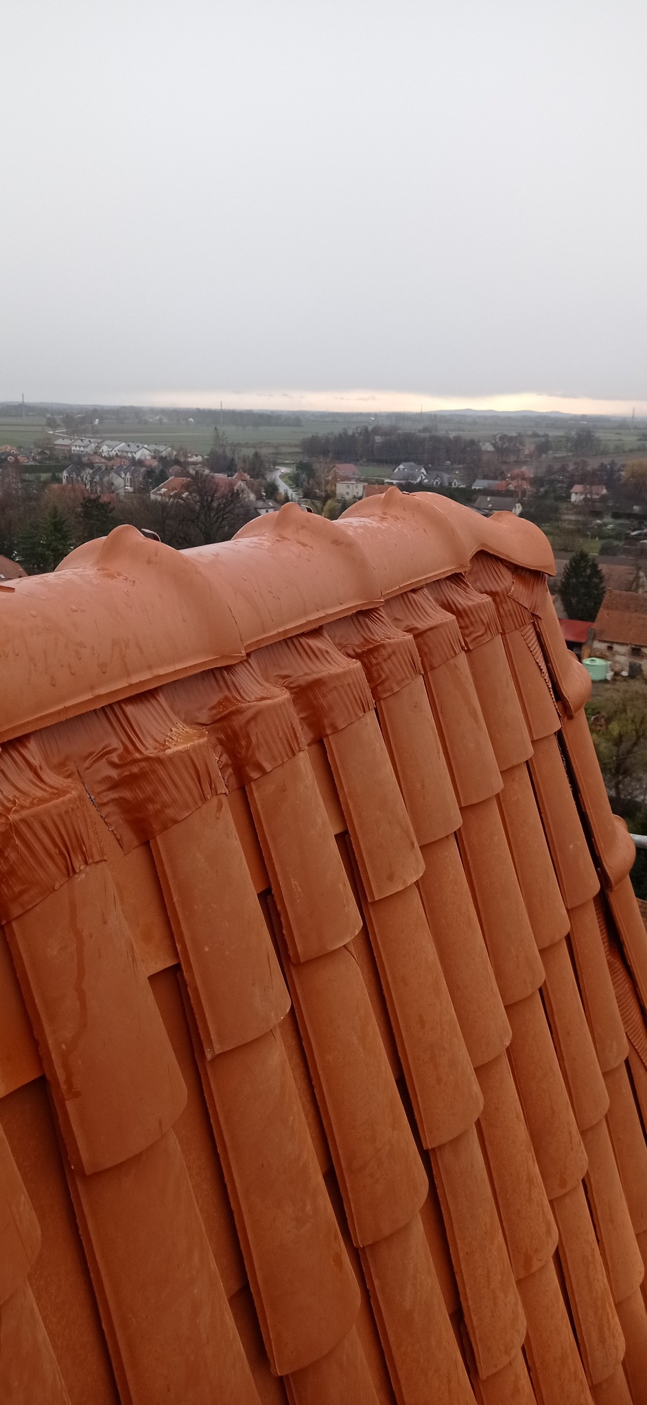 Czerwona dachówka ceramiczna na dachu domu w pochmurny dzień, widok z góry na fragment pokrycia z widocznymi elementami mocującymi, w tle zabudowa mieszkalna.