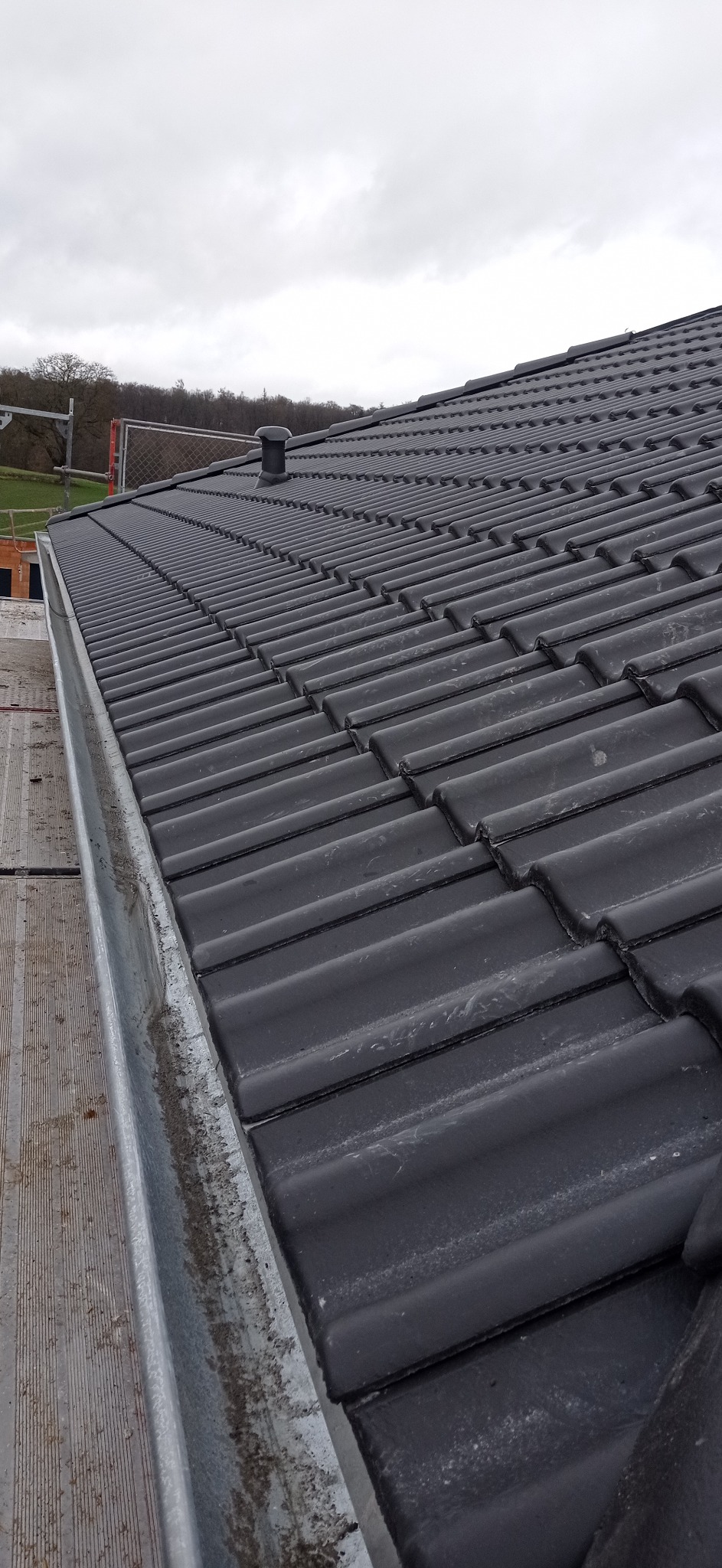 Nowe, ciemnoszare dachówki ceramiczne ułożone na dachu, widoczna metalowa rynna i fragment rusztowania, pochmurne niebo w tle.
