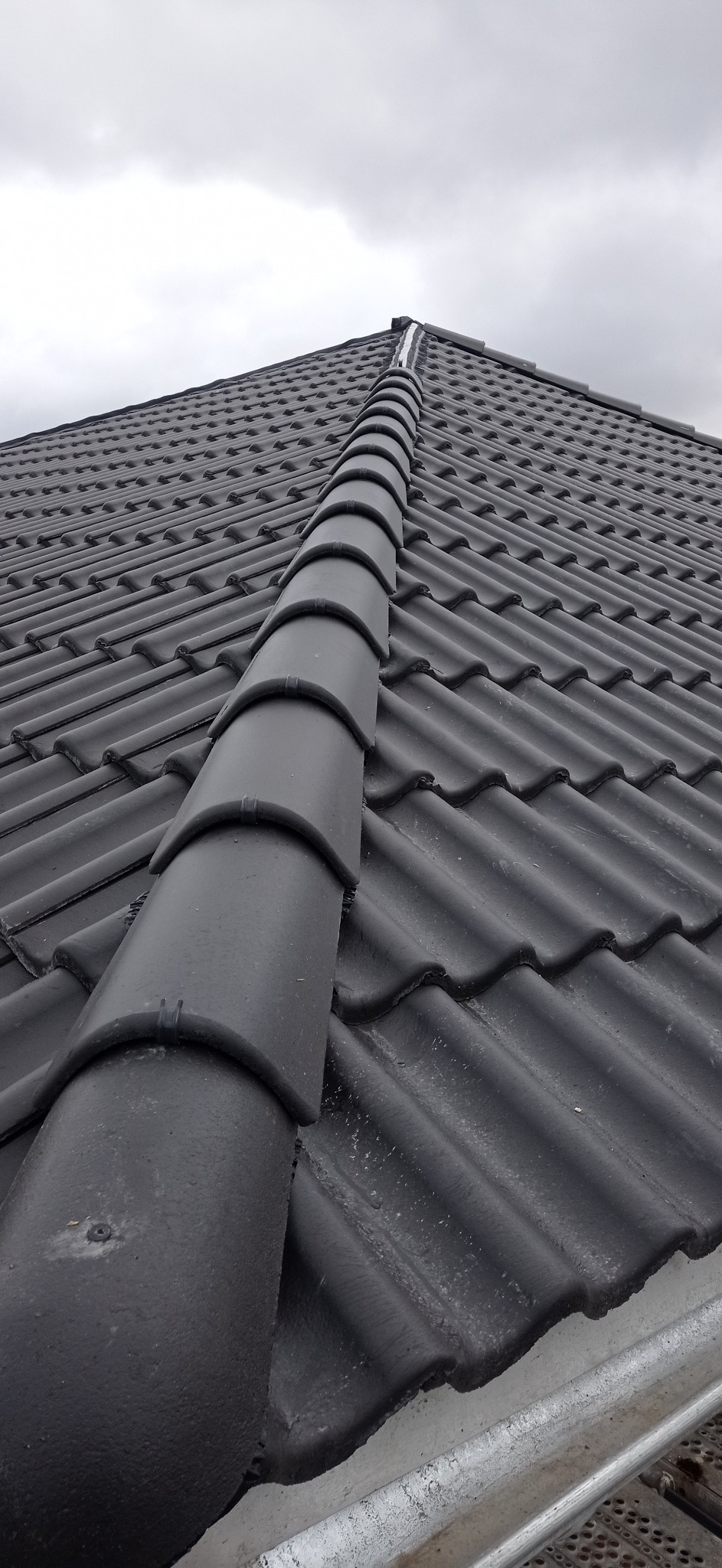 Nowy dach z ciemnej dachówki ceramicznej, widok z dołu na kalenicę i rynny, pochmurne niebo w tle.