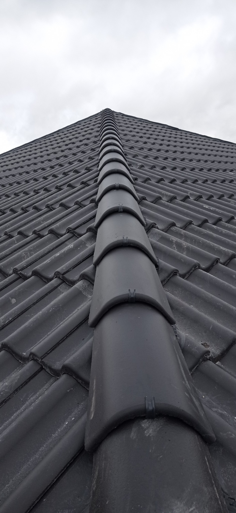 Szczegółowe ujęcie czarnych, ceramicznych dachówek karpiówek ułożonych na dachu, z widocznym kalenicą, na tle pochmurnego nieba.