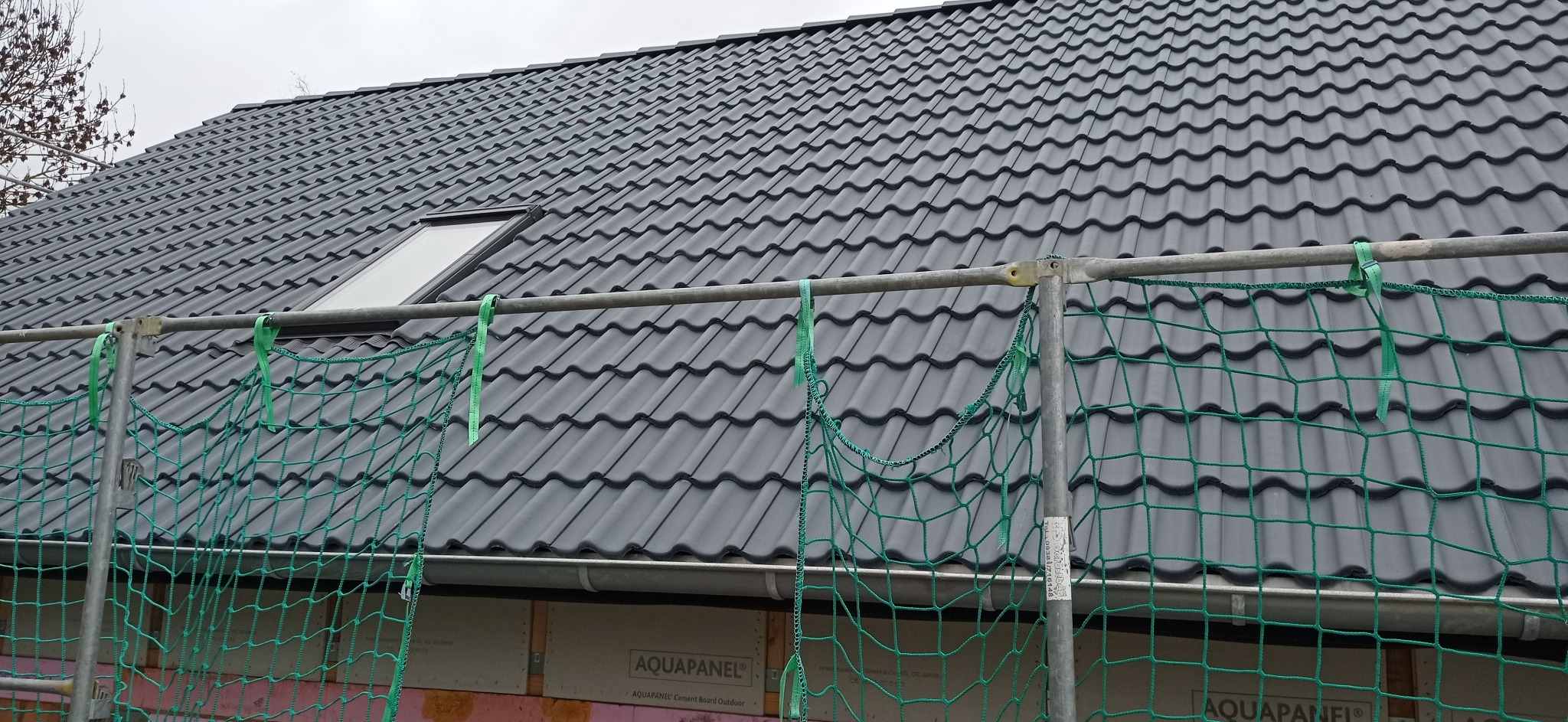 Nowy dach z szarej dachówki ceramicznej z oknem dachowym, widoczny przez siatkę zabezpieczającą na rusztowaniu.