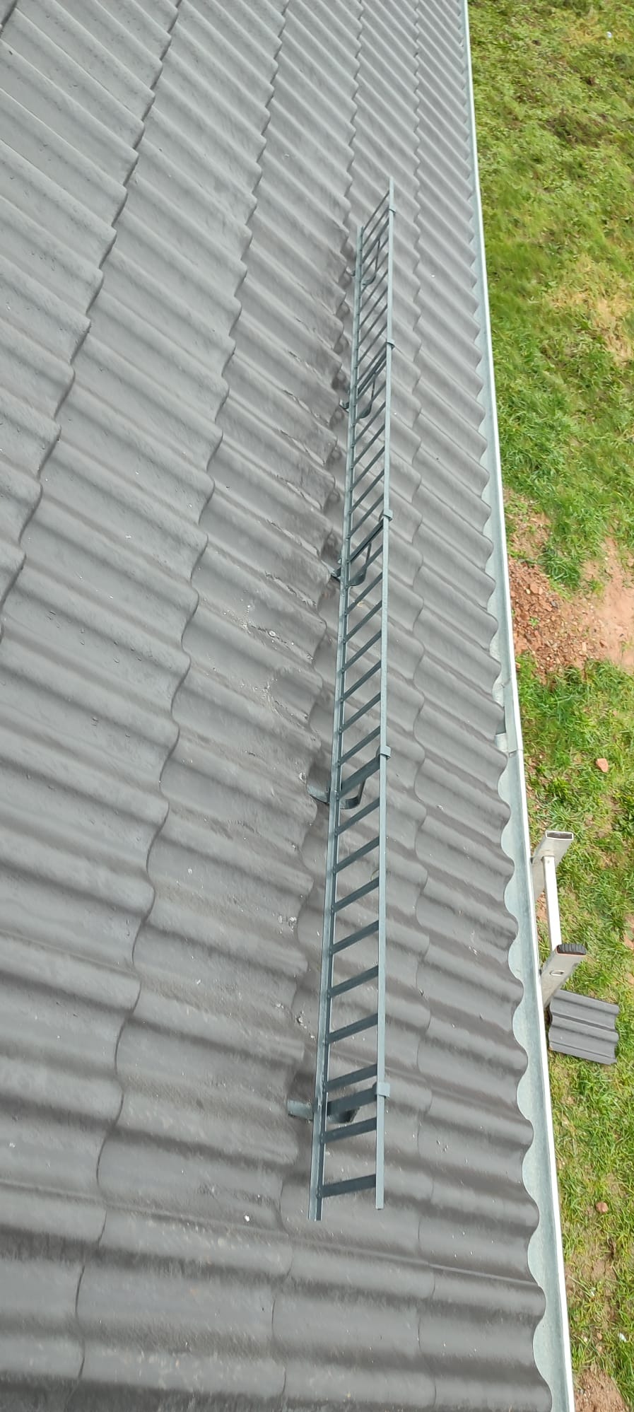 Szare dachówki z zamontowanym stalowym stopniem kominiarskim i widoczną rynną, na tle zielonej trawy.