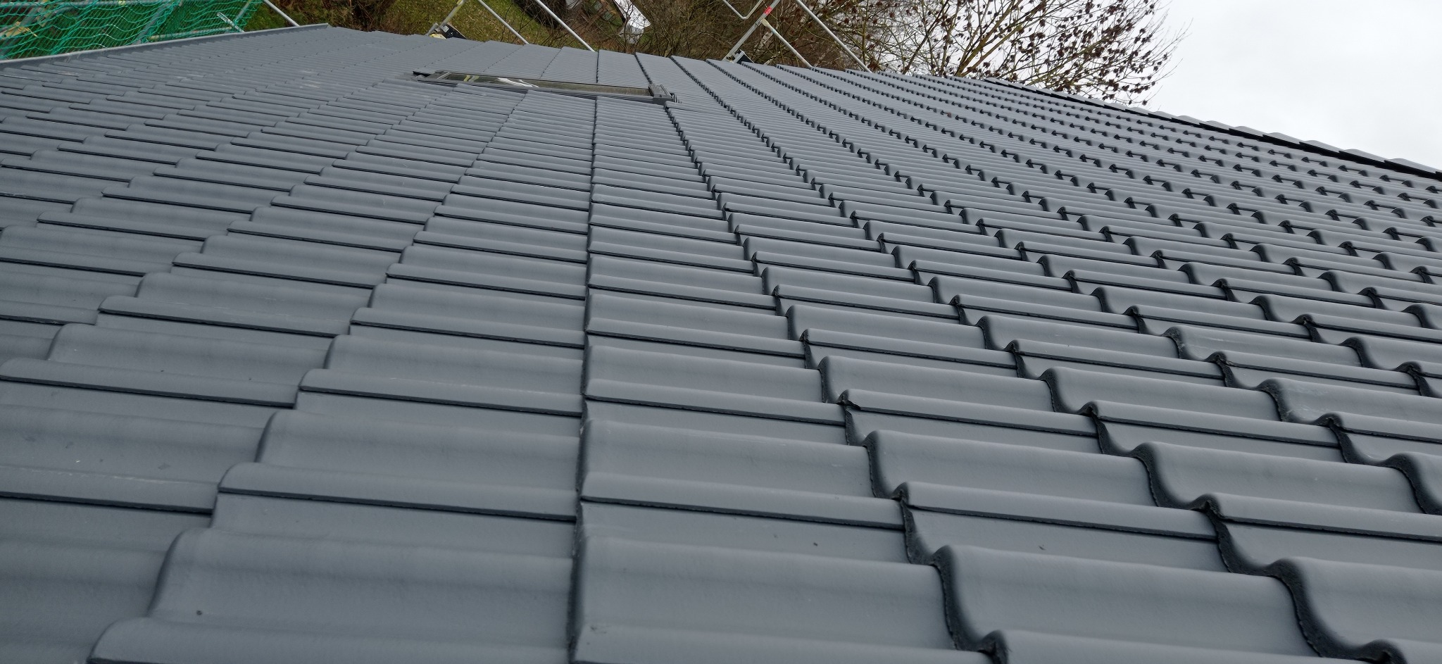 Nowy, szary dach z dachówką ceramiczną, widoczne okno dachowe i rusztowanie w tle.