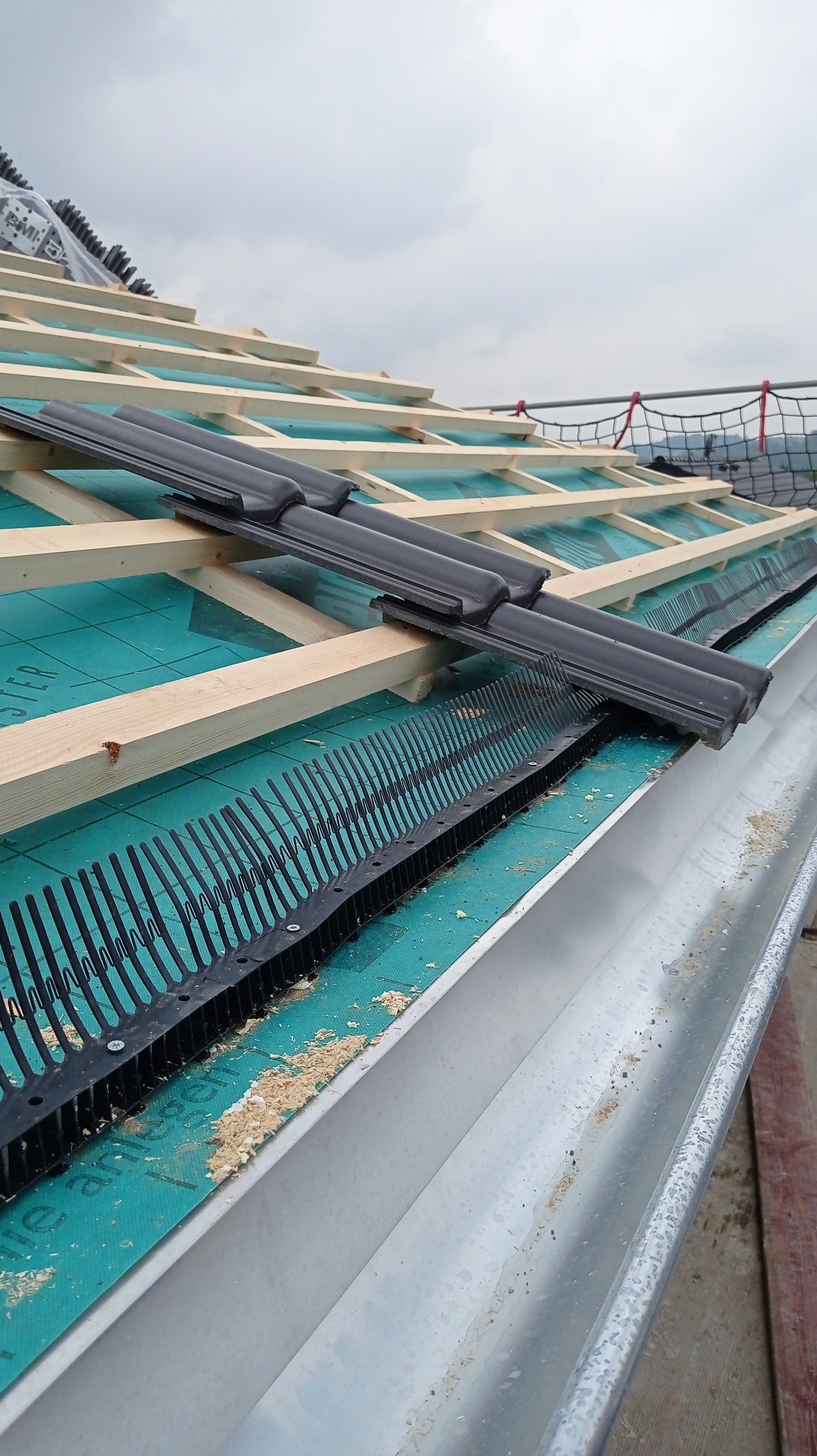 Montaż zabezpieczeń przed ptakami na dachu z widoczną konstrukcją drewnianą i rynną, fragmenty trocin na membranie dachowej.