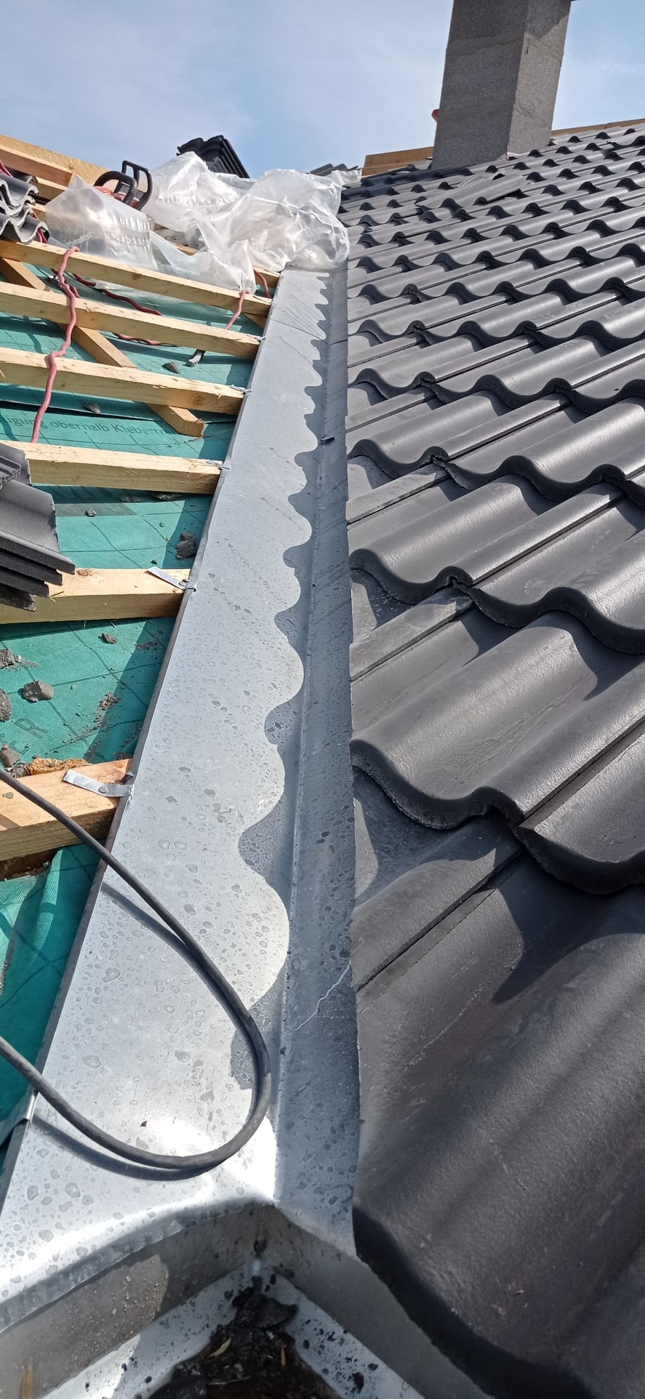 Fragment dachu w trakcie budowy: widoczne drewniane elementy konstrukcyjne, zielona membrana, zamontowany metalowy pas przy krawędzi oraz ciemnoszare dachówki karpiówki.