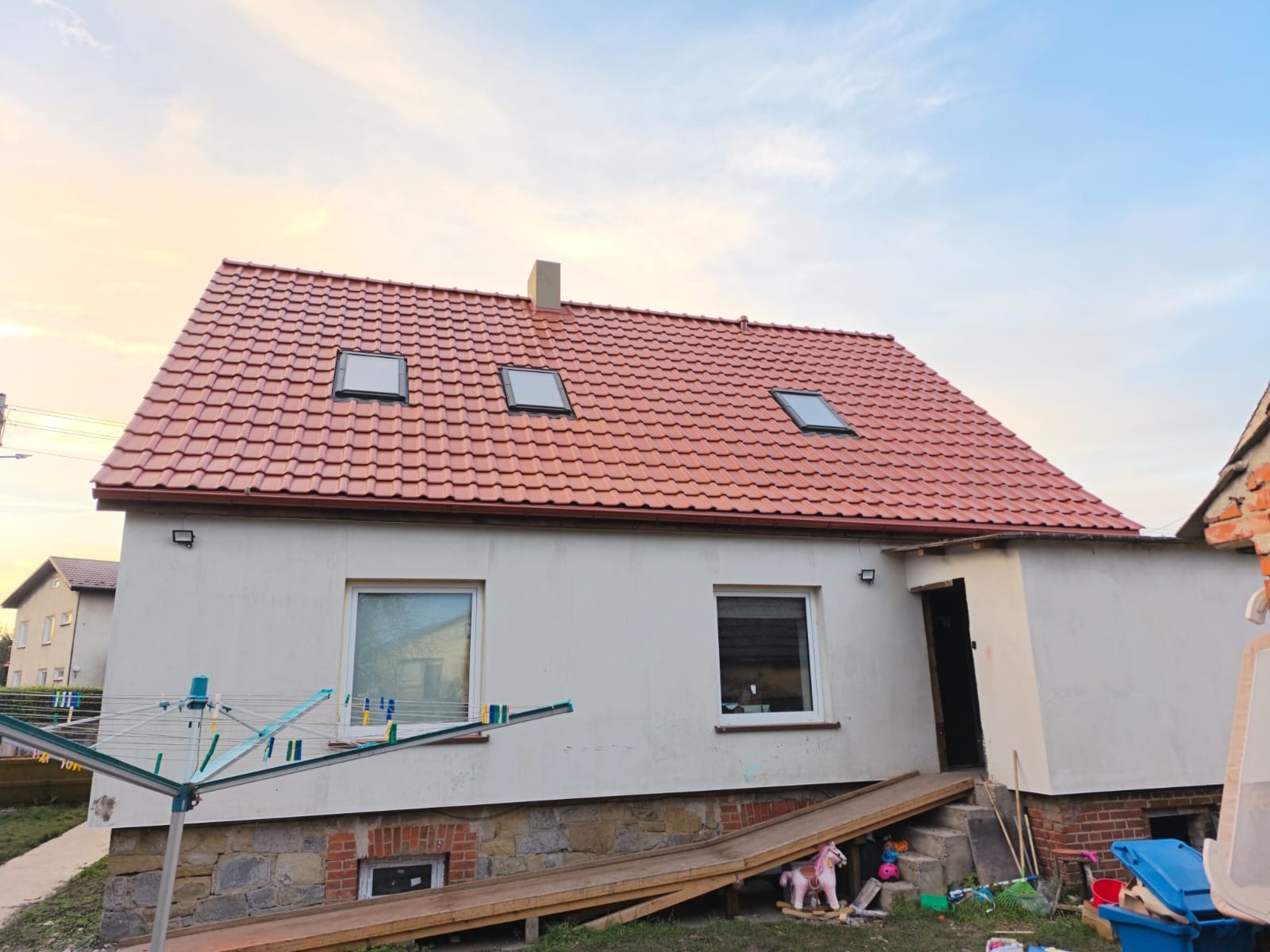 Dom z nowym, czerwonym dachem z dachówki ceramicznej i trzema oknami dachowymi. Widoczne rynny i fragment elewacji. Ujęcie z poziomu gruntu, w tle błękitne niebo.