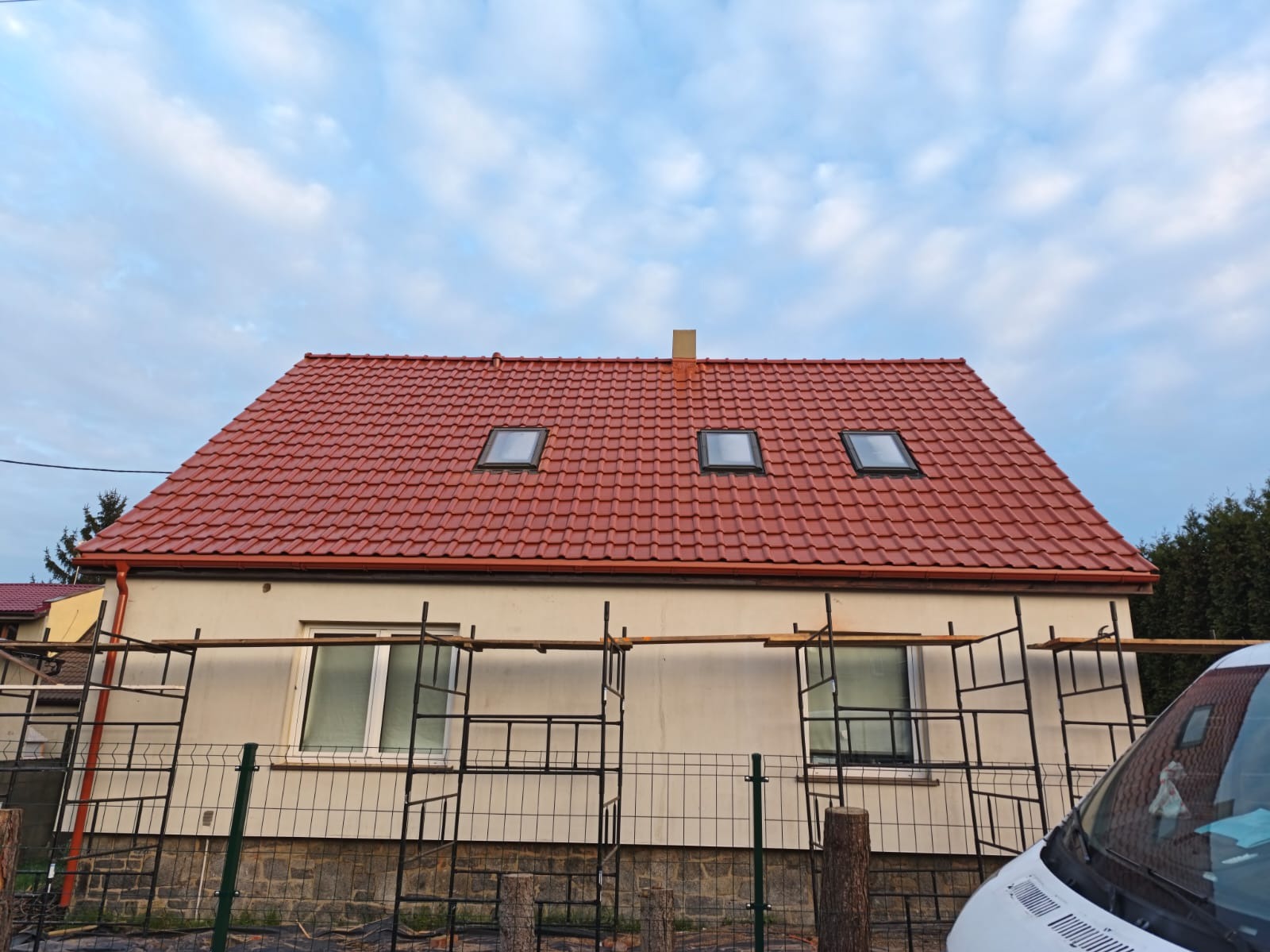Dom z nowym, czerwonym dachem z trzema oknami dachowymi. Widoczne rusztowania i fragment samochodu. Ujęcie z dołu, podkreślające świeżość pokrycia.