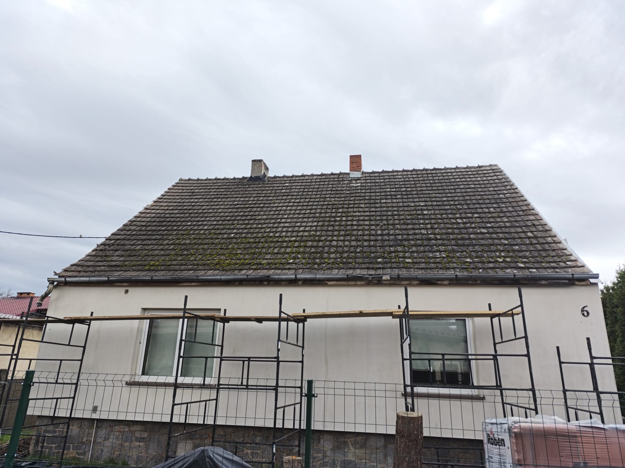 Dom jednorodzinny z szarym, zarośniętym mchem dachem, częściowo osłonięty rusztowaniem. Widoczny fragment ogrodzenia i paleta z dachówkami. Pochmurne niebo w tle.