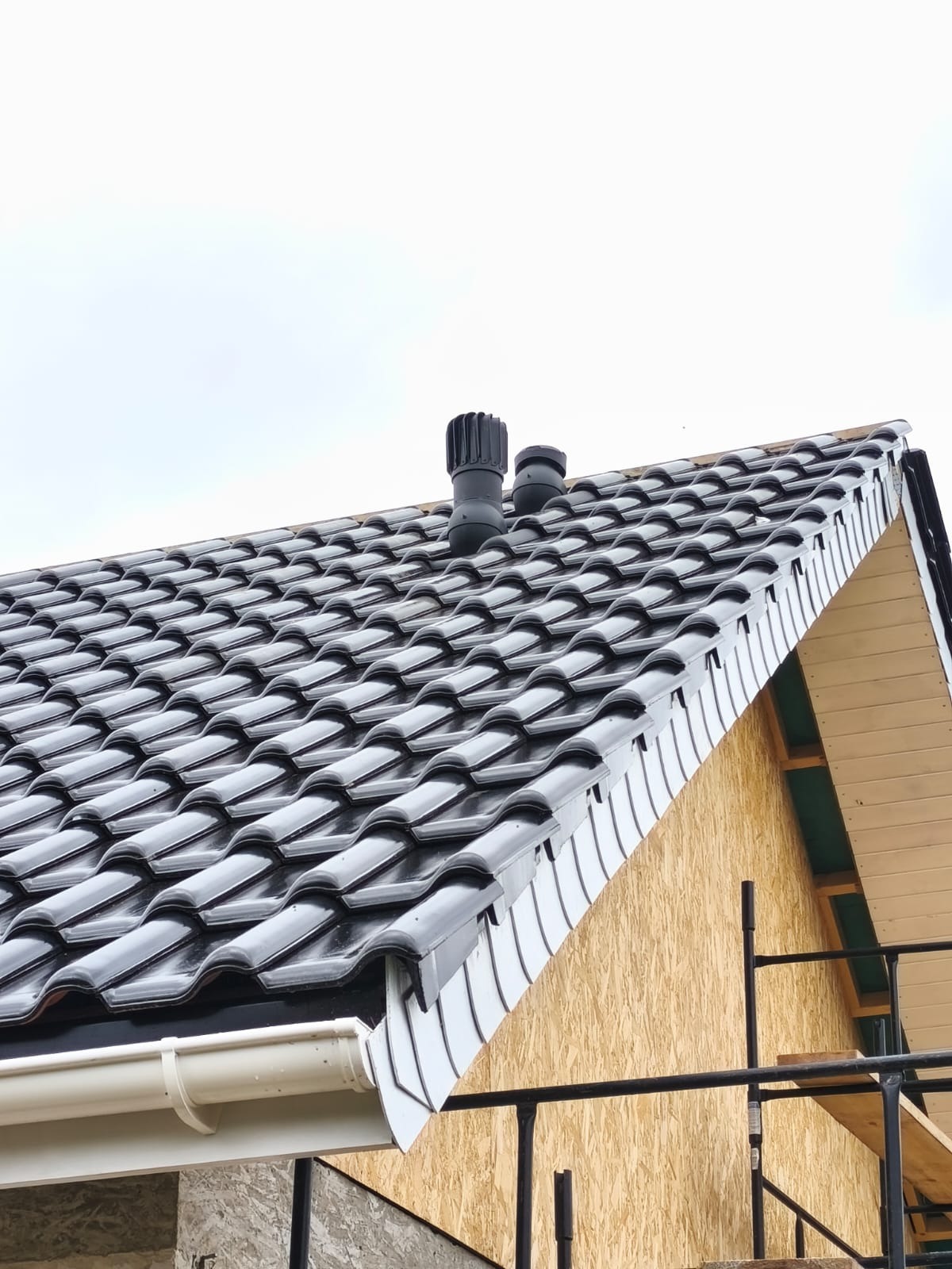 Czarny dach z dachówki ceramicznej z dwoma kominkami wentylacyjnymi, białą rynną i fragmentem rusztowania na tle jasnego nieba.