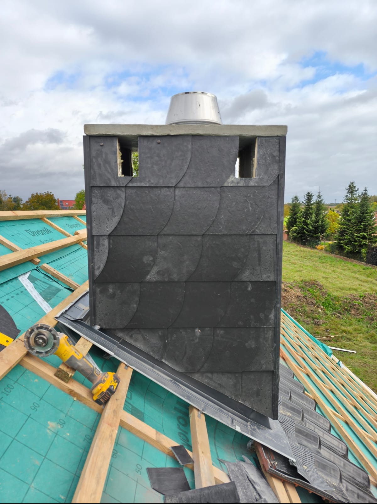 Kominek obłożony łupkiem na dachu w trakcie budowy, widoczne narzędzia i membrana dachowa. Ujęcie z bliska, podkreślające detale wykończenia.