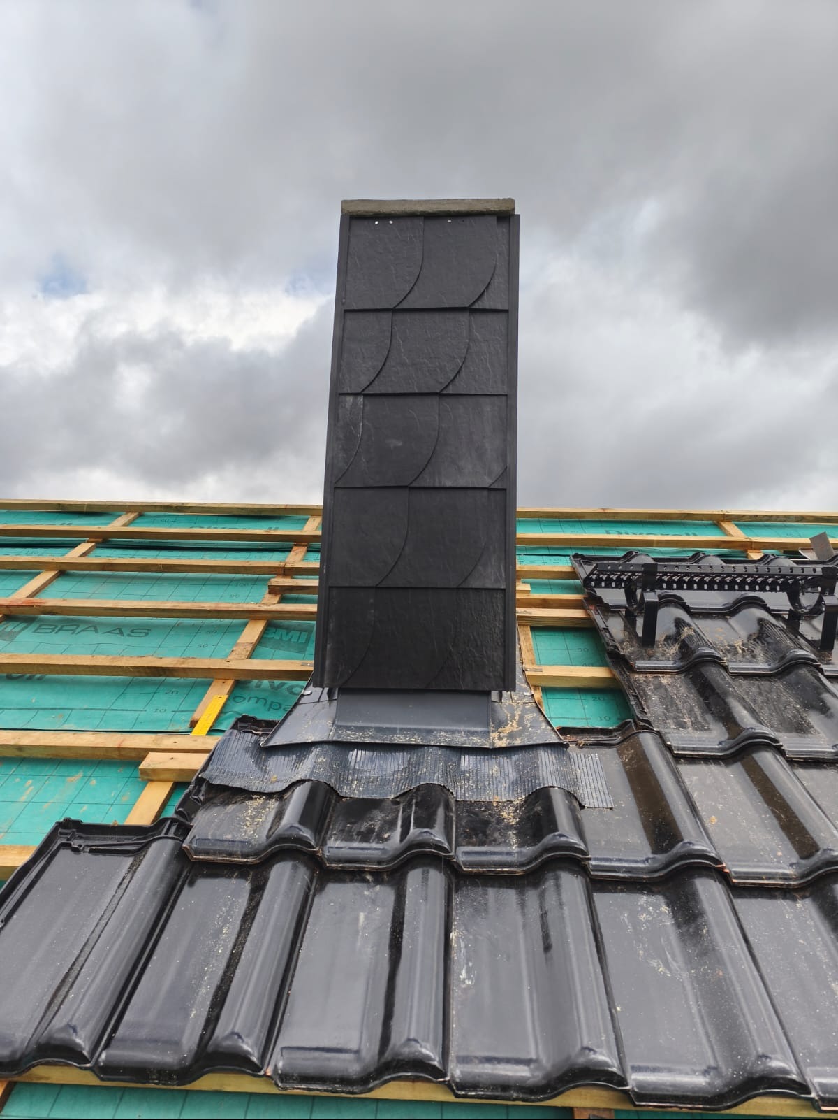 Fragment dachu z czarną dachówką ceramiczną i kominem obłożonym łupkiem, widoczne elementy konstrukcji drewnianej i membrany dachowej na tle pochmurnego nieba.