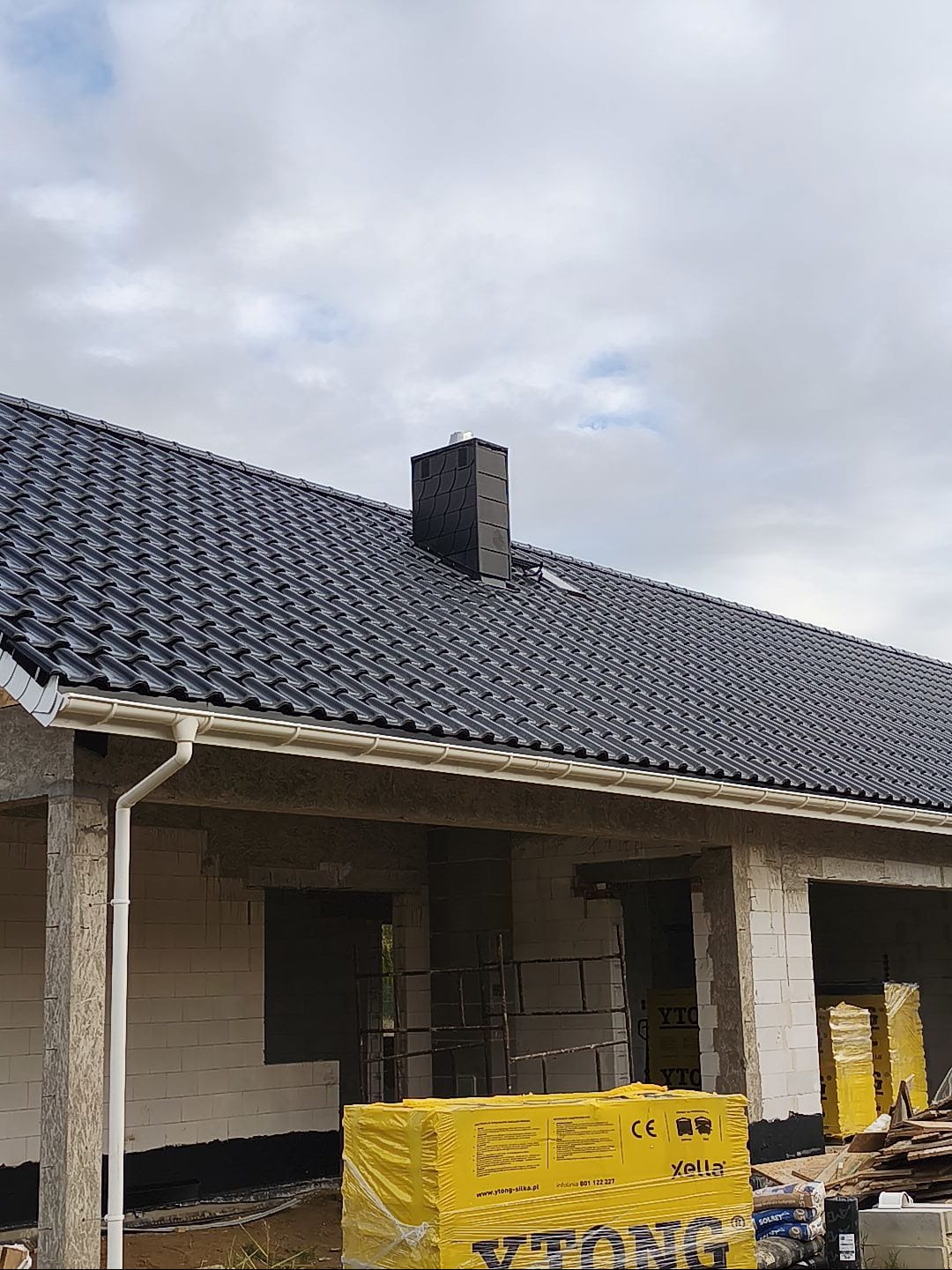 Nowy, czarny dach z ceramicznej dachówki karpiówki i jasne rynny na surowym budynku z betonu komórkowego. Widoczny komin i zapakowane bloczki Ytong.