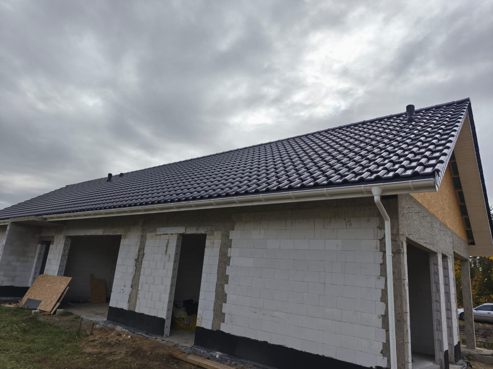 Nowy, czarny dach z rynnami na budynku w trakcie budowy, widok z dołu na tle pochmurnego nieba. Białe ściany z betonu komórkowego, widoczne otwory okienne i drzwiowe.