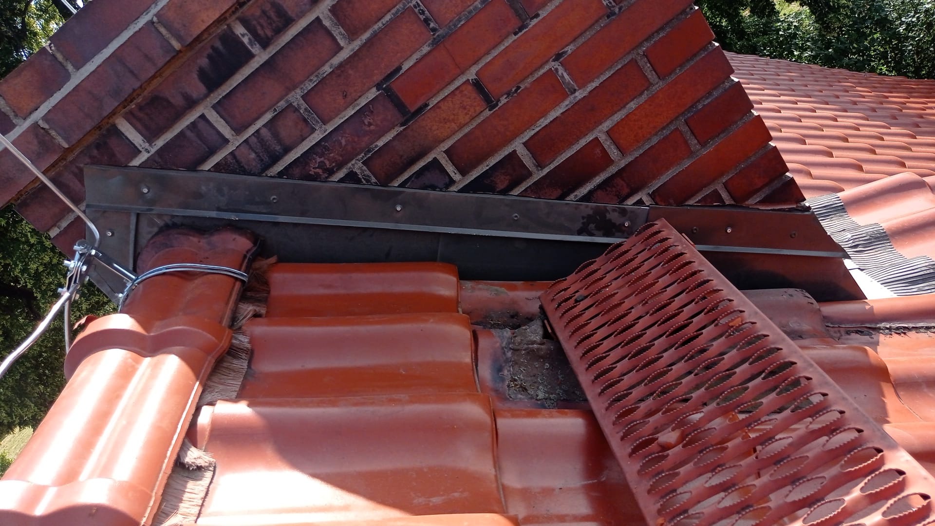 Ceglany komin z metalowym obramowaniem, dach z czerwonej dachówki ceramicznej, zabezpieczenie rynny przed liśćmi. Widok z góry na fragment dachu.