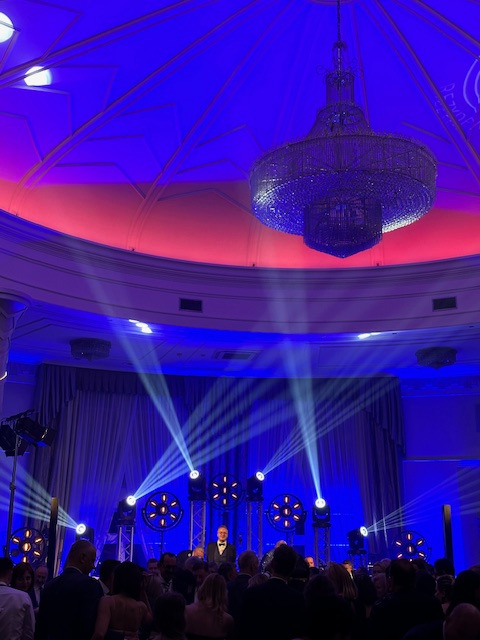 Sala balowa z oświetleniem scenicznym, kryształowym żyrandolem i osobą na scenie w smokingu, widziana z perspektywy tłumu.