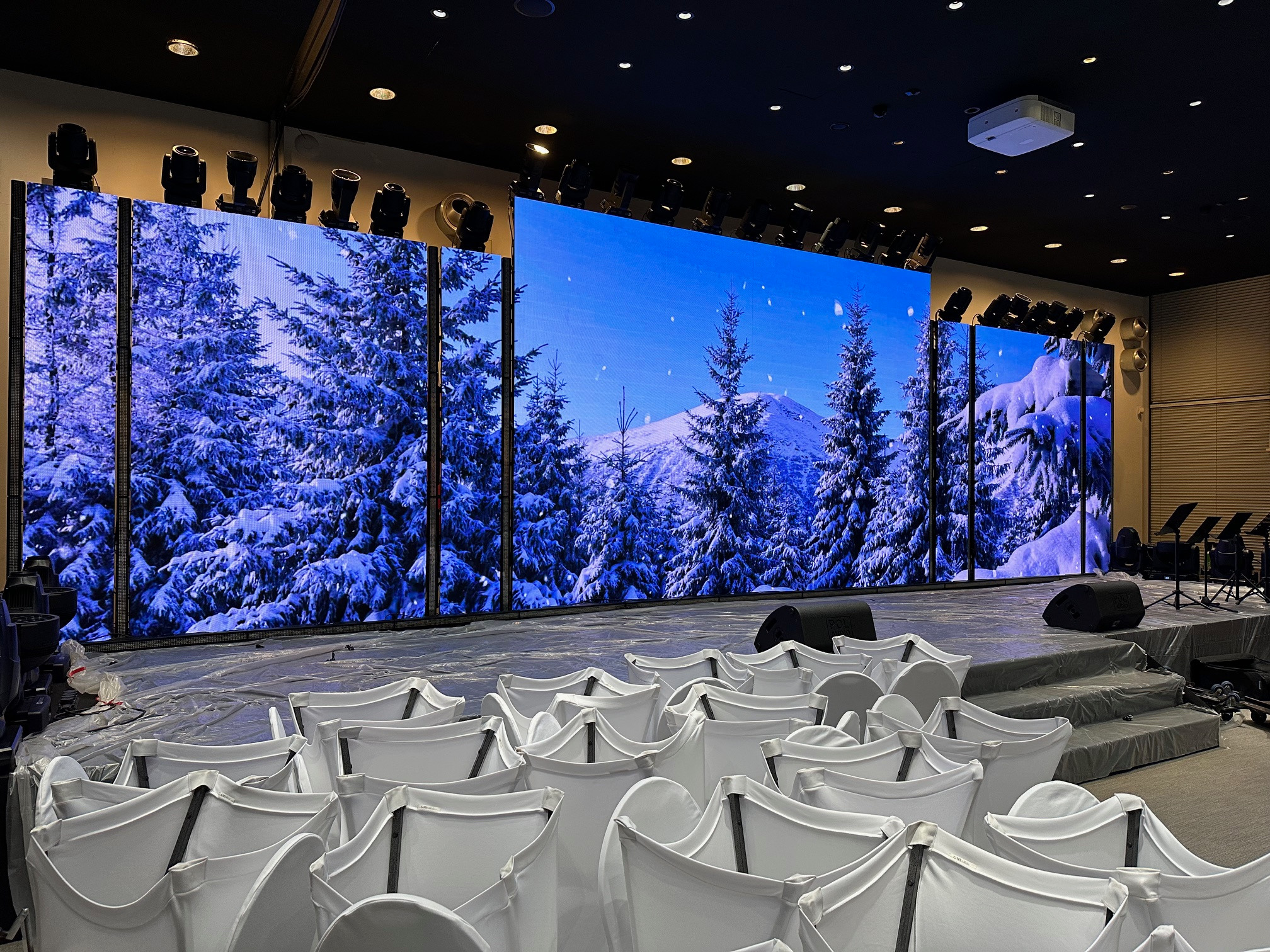 Scena przed wydarzeniem: duży ekran LED wyświetla zimowy krajobraz z zasypanymi śniegiem drzewami i górami, oświetlenie sceniczne na górze, rzędy białych krzeseł z pokrowcami na widowni...