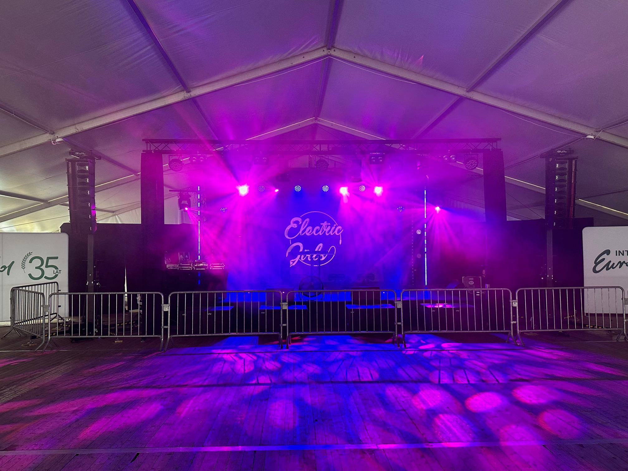 Scena oświetlona fioletowo-niebieskim światłem, widoczne nagłośnienie, barierki ochronne i logo zespołu Electric Girls.