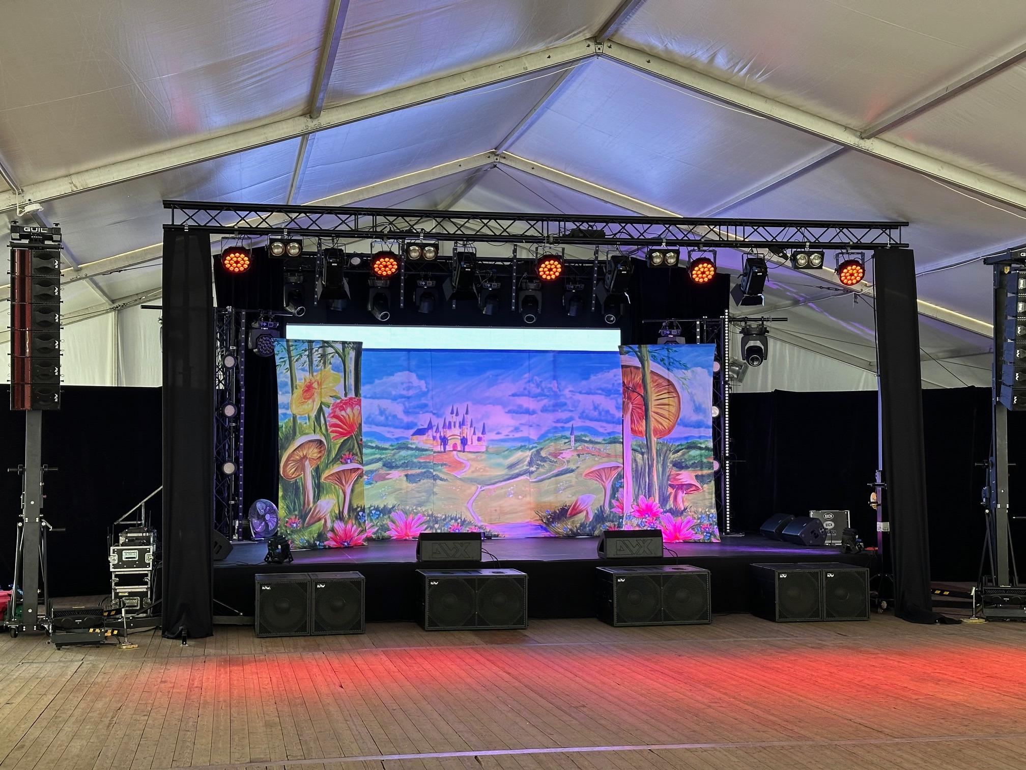 Scena z nagłośnieniem i oświetleniem w namiocie, dekorowana bajkowym tłem z motywem zamku i grzybów, liczne głośniki i reflektory.