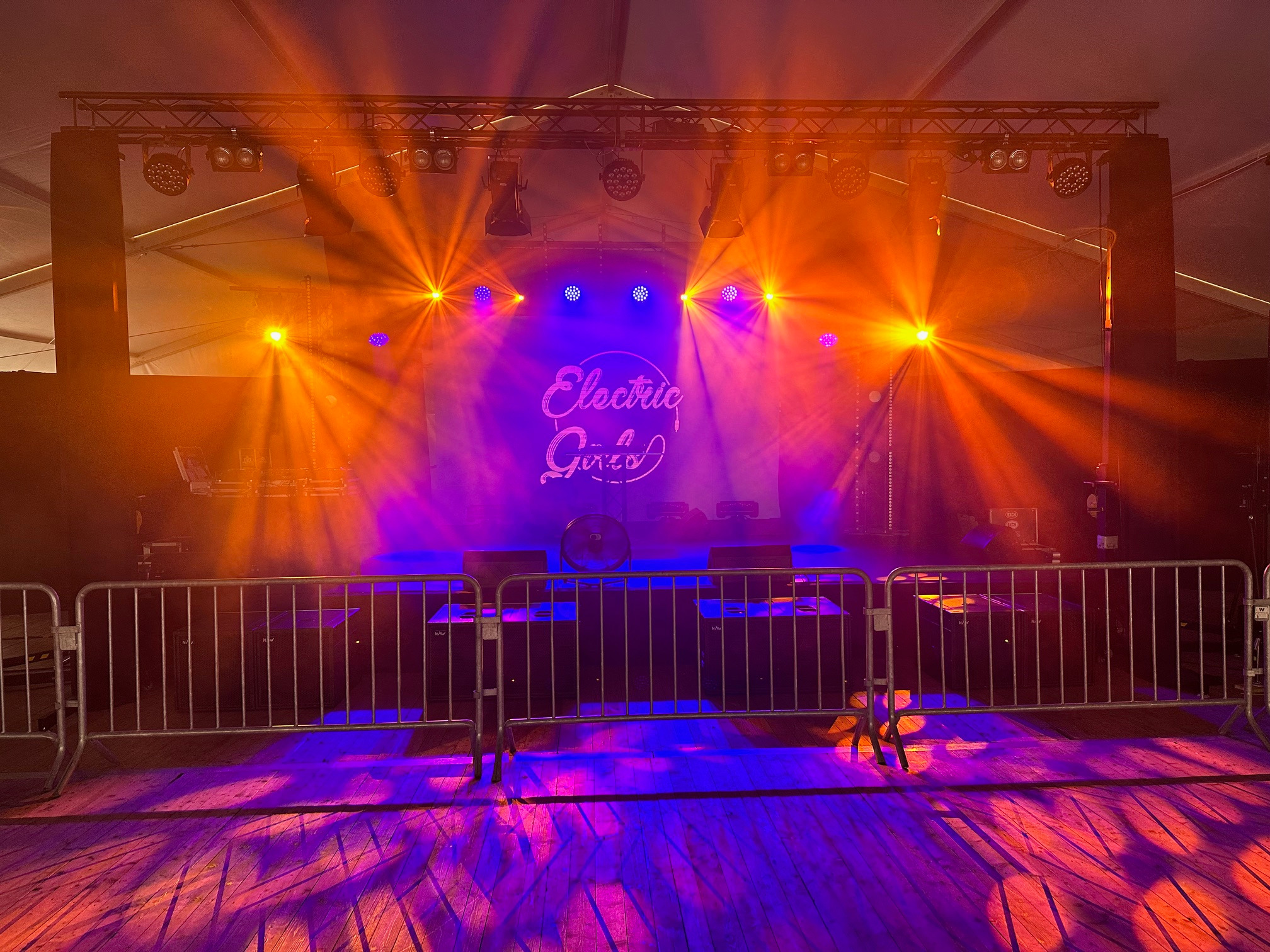Scena oświetlona pomarańczowymi i niebieskimi światłami, z widocznym nagłośnieniem i metalową barierką na pierwszym planie, podłoga z drewnianych desek.