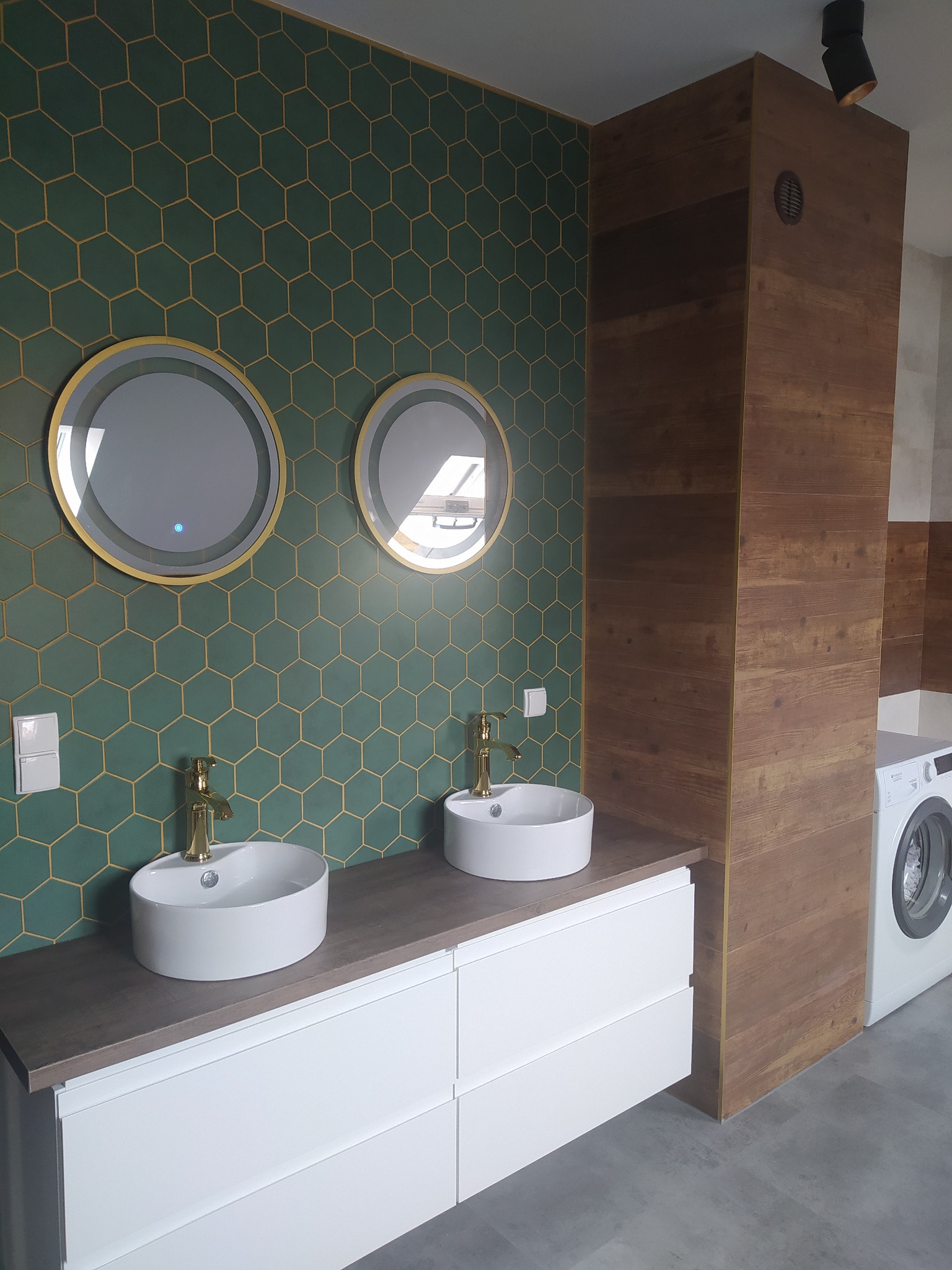 Nowoczesna łazienka z zielonymi heksagonalnymi płytkami, okrągłymi lustrami z podświetleniem i białymi umywalkami nablatowymi z złotą armaturą. Widoczna pralka.