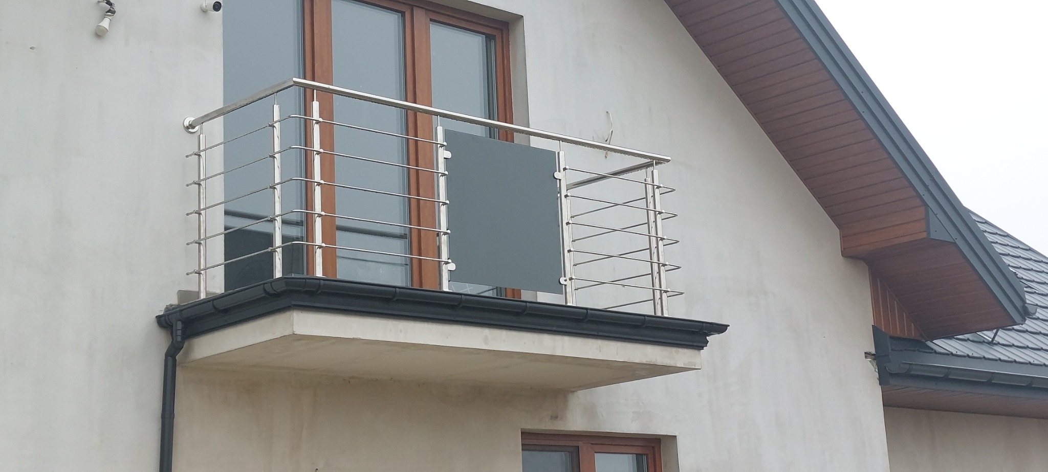 Balkon z poziomą, metalową balustradą z elementami szarego szkła, zamontowany przy budynku z drewnianą podbitką.