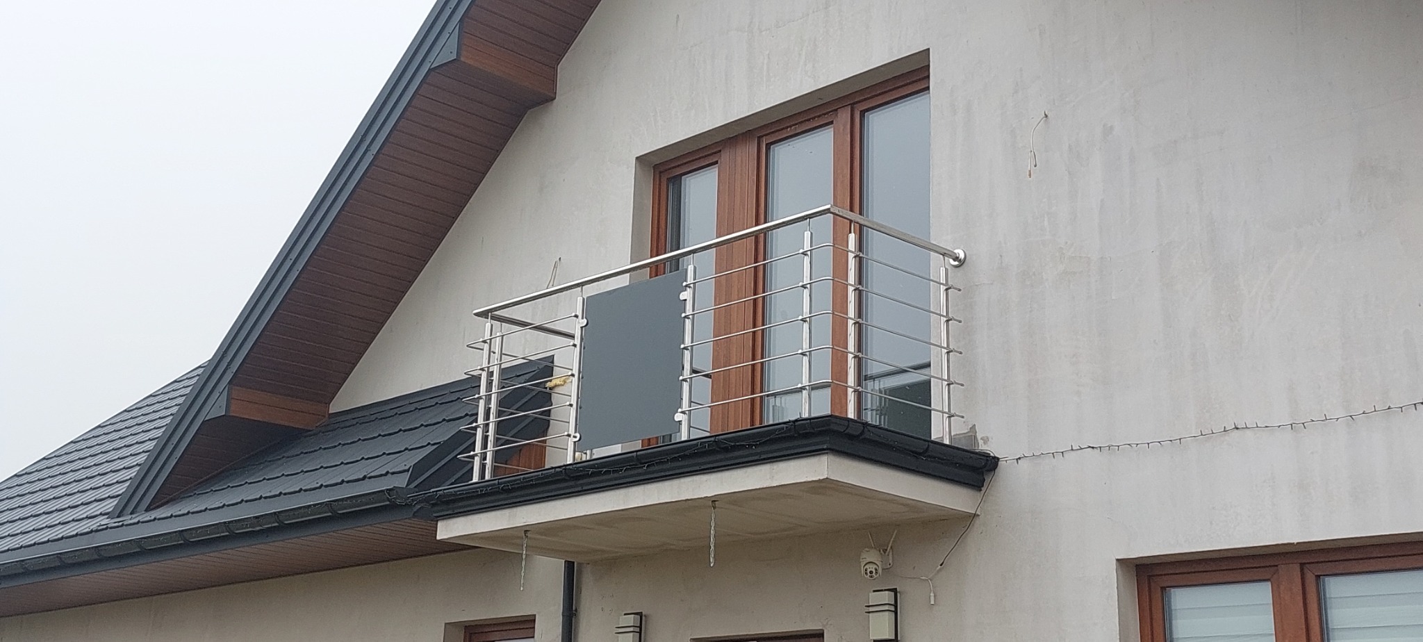 Balkon z balustradą nierdzewną z pionowymi szczeblami i szarą wstawką, zamontowany przy elewacji domu z drewnianą stolarką okienną i dachem z ciemnej dachówki.