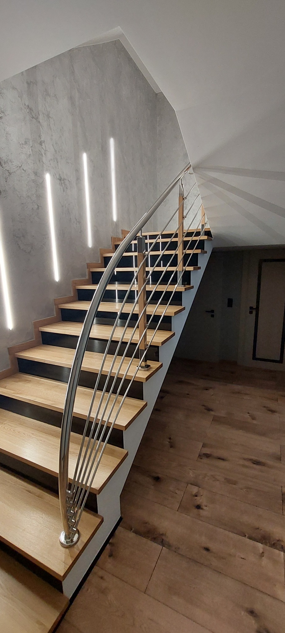 Nowoczesne schody z drewnianymi stopniami, czarnymi podstopnicami i zaokrągloną, wieloliniową balustradą ze stali nierdzewnej, oświetlone pionowymi lampami ściennymi.