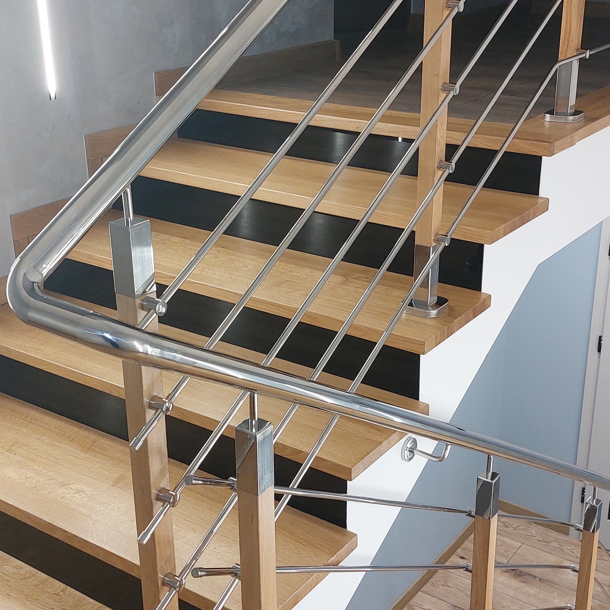 Schody drewniane z nowoczesną, polerowaną balustradą ze stali nierdzewnej z drewnianymi słupkami, widok z dołu na górę, z elementami czerni na podstopnicach.