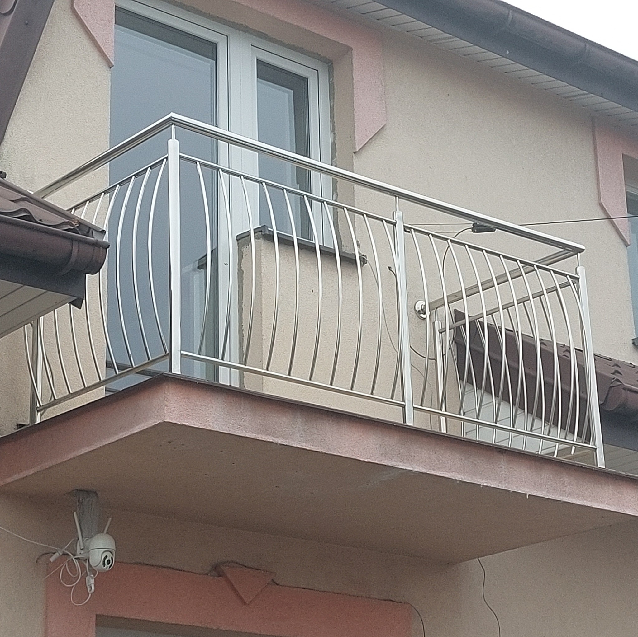 Balkon z nowoczesną, giętą, metalową barierką, widoczna kamera monitoringu pod balkonem.