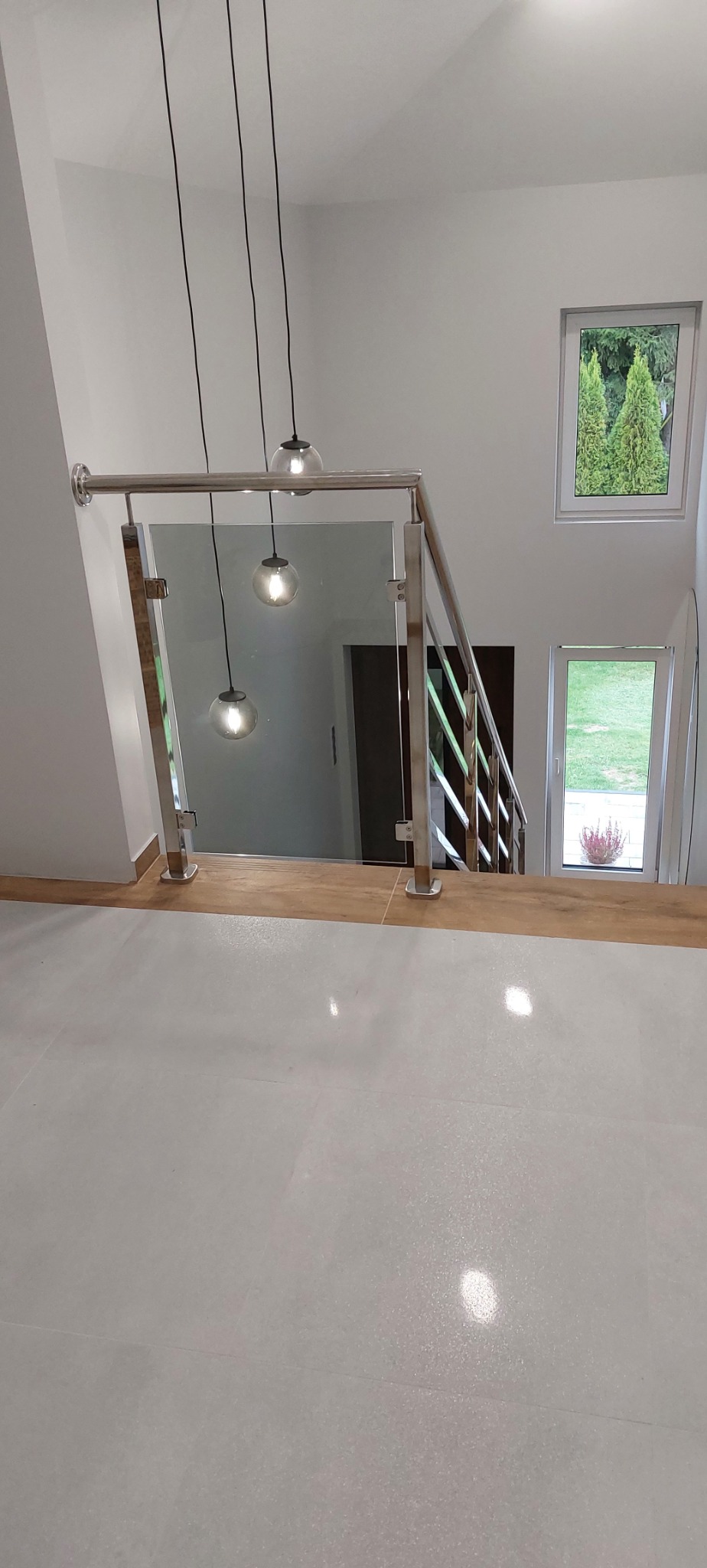 Szklana balustrada ze słupkami ze stali na schodach w nowoczesnym domu, widok z góry na białe płytki podłogowe i wiszące lampy z kulistymi kloszami.