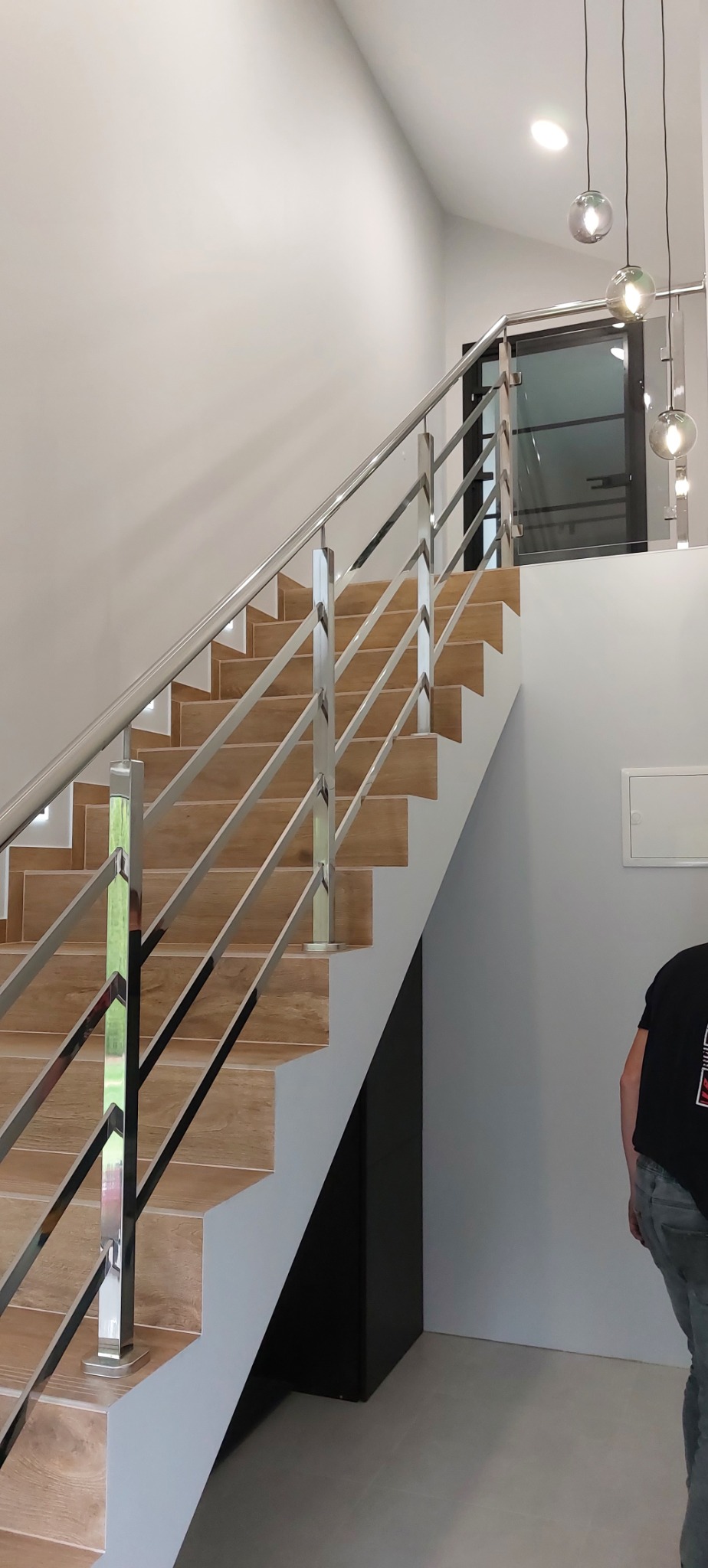 Wnętrze domu ze schodami wyłożonymi płytkami imitującymi drewno, zabezpieczonymi prostą, nowoczesną balustradą ze stali nierdzewnej z wypełnieniem z poziomych prętów, widoczne wiszące lampy...