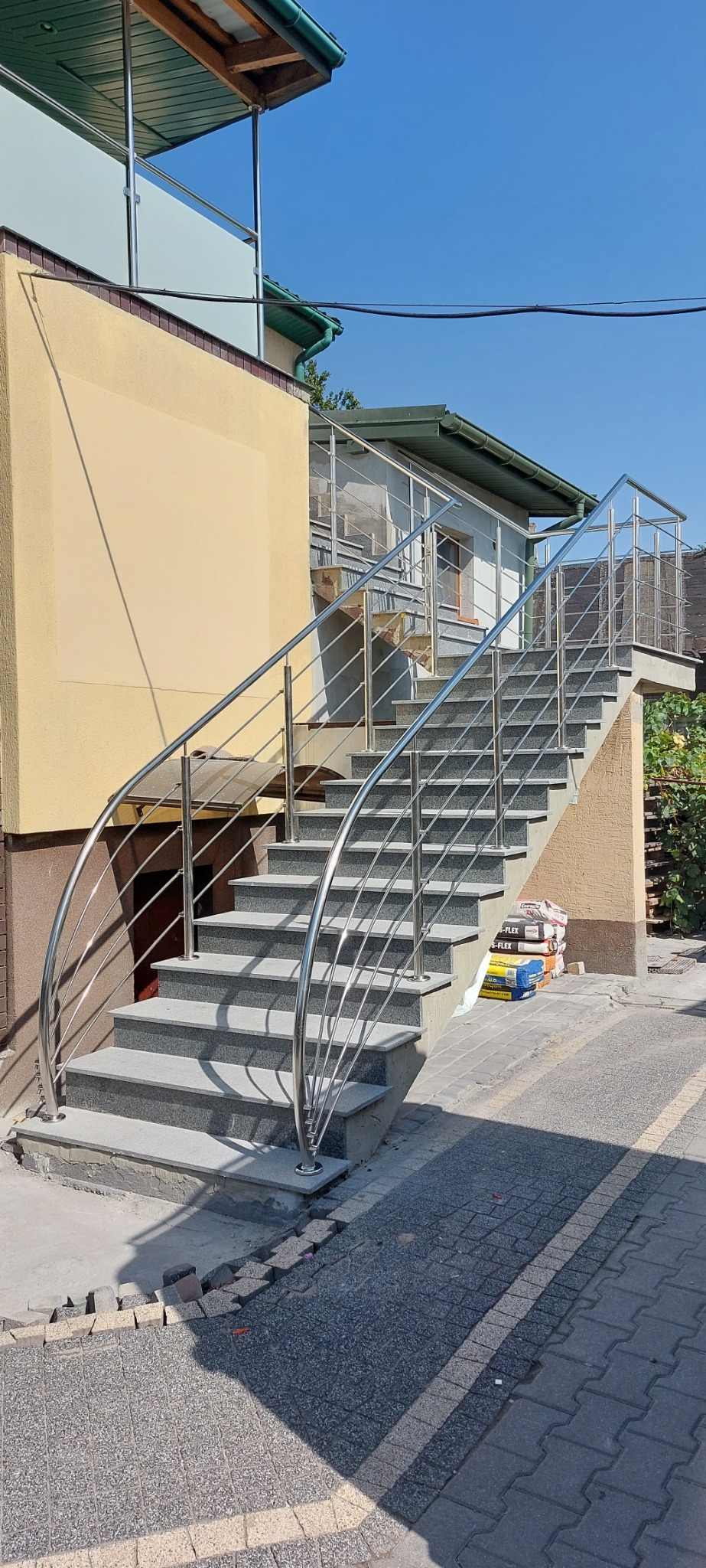 Zewnętrzne schody z szarego granitu prowadzące do budynku, zabezpieczone balustradą z polerowanej stali nierdzewnej o zaokrąglonym kształcie, widoczne worki z zaprawą budowlaną.