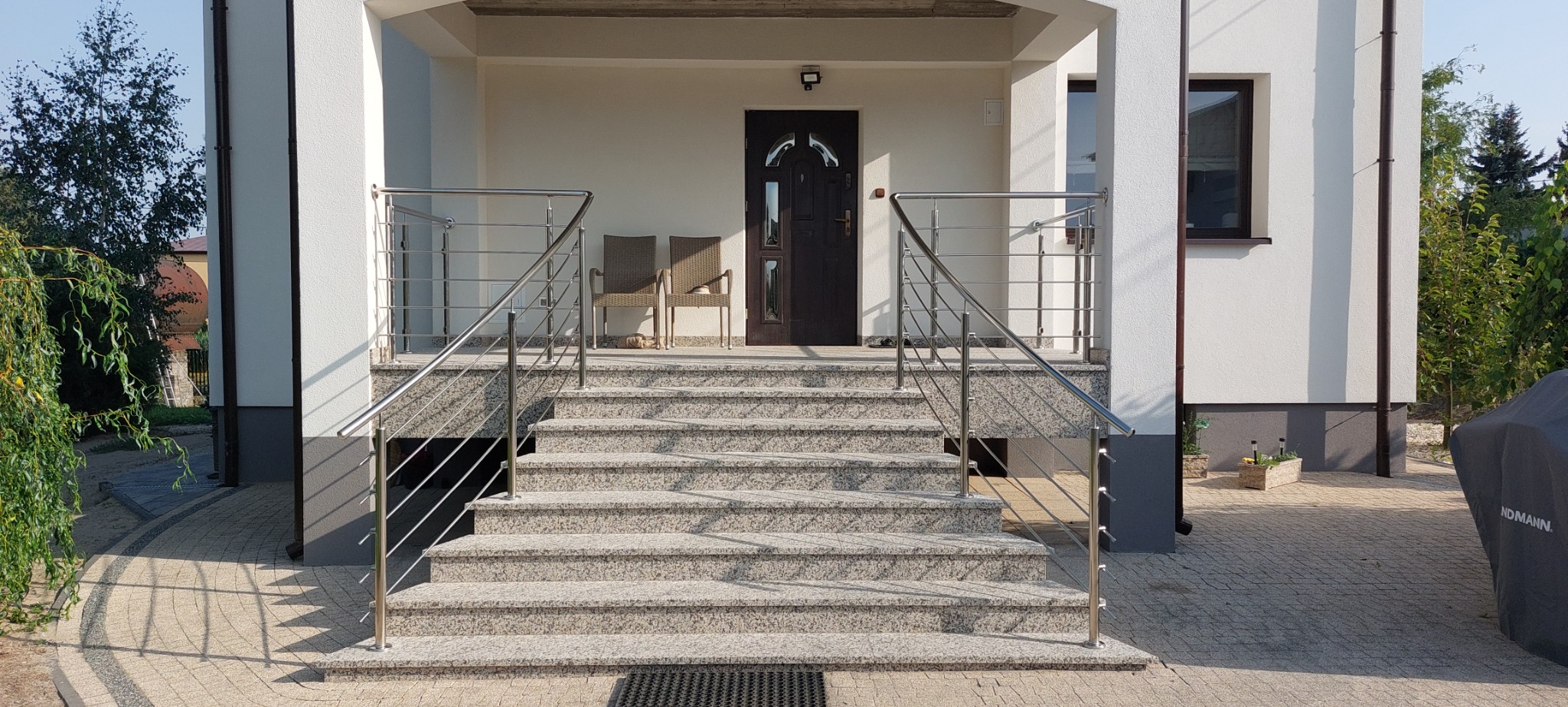 Zewnętrzne schody wejściowe do domu wyłożone jasnym granitem, zabezpieczone prostą, nowoczesną balustradą z poziomych, cienkich rurek.