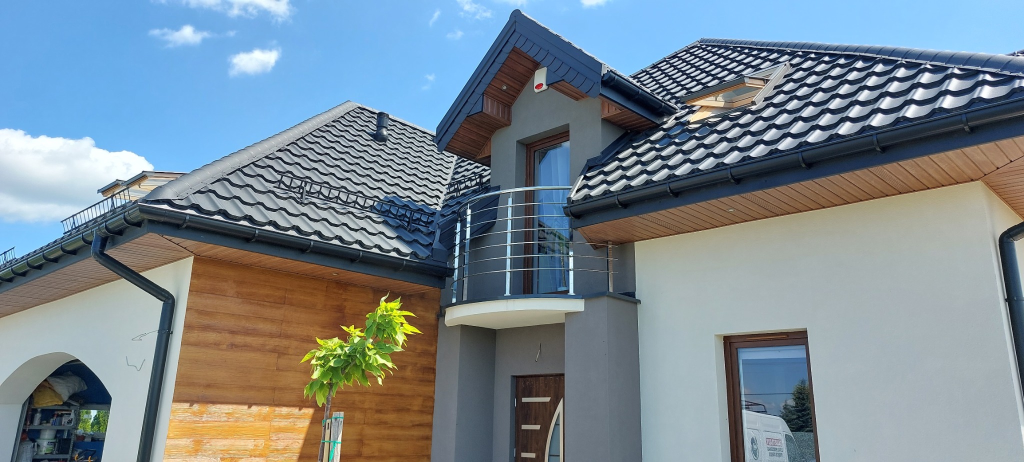 Nowoczesny dom jednorodzinny z balkonem zabezpieczonym okrągłą balustradą ze stali, czarnym dachem i elementami drewnianymi na elewacji w słoneczny dzień.